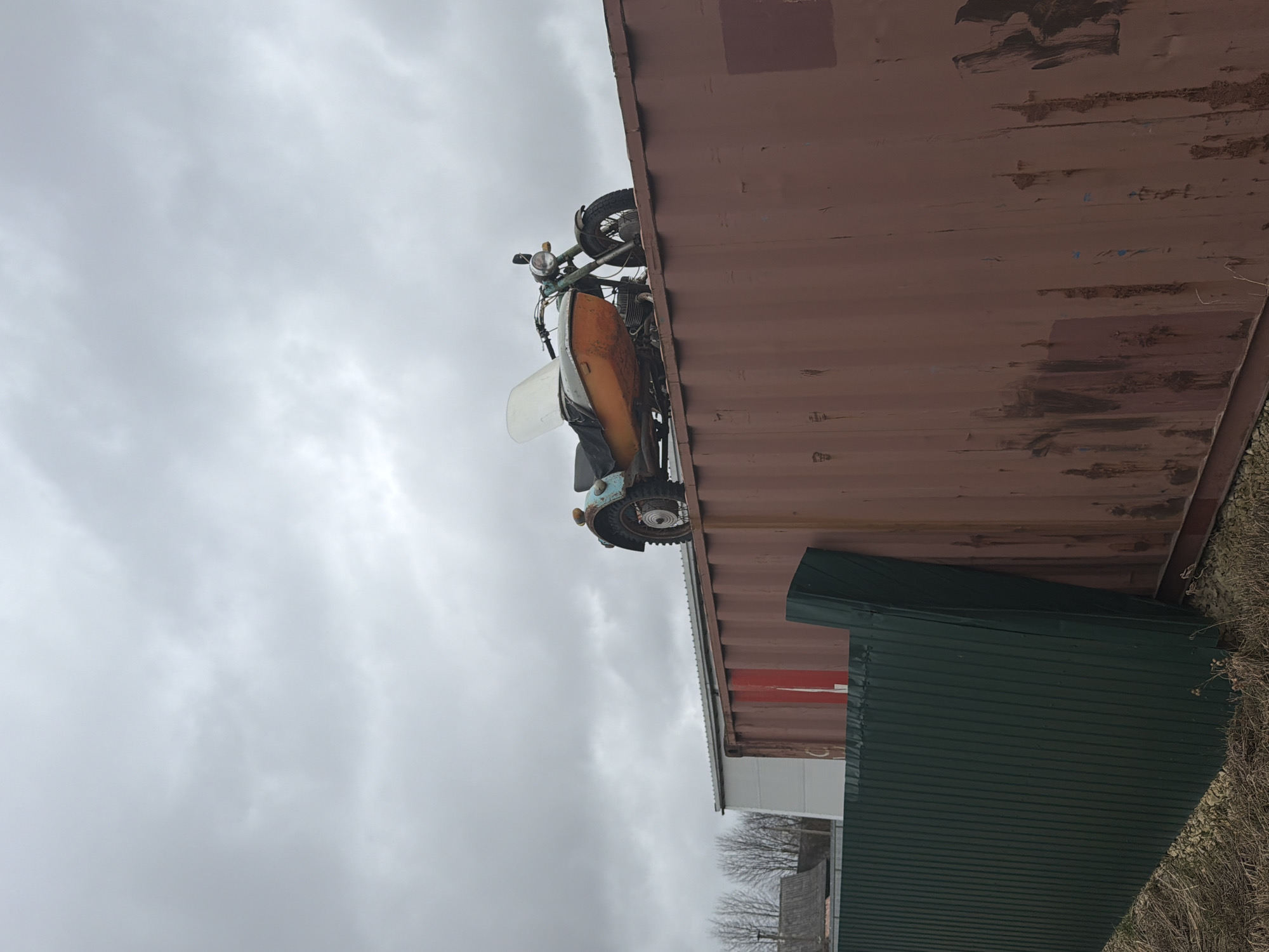 The image depicts a vintage motorcycle mounted on the side of a brown shipping container. The motorcycle appears to be a light-colored vintage model with an orange and white sidecar attached. The container is positioned near a smaller structure covered with green siding. The sky is overcast, and the surrounding environment includes bare trees and foliage, suggesting a late fall or winter scene. The photo was taken in Torosovo, Russia.