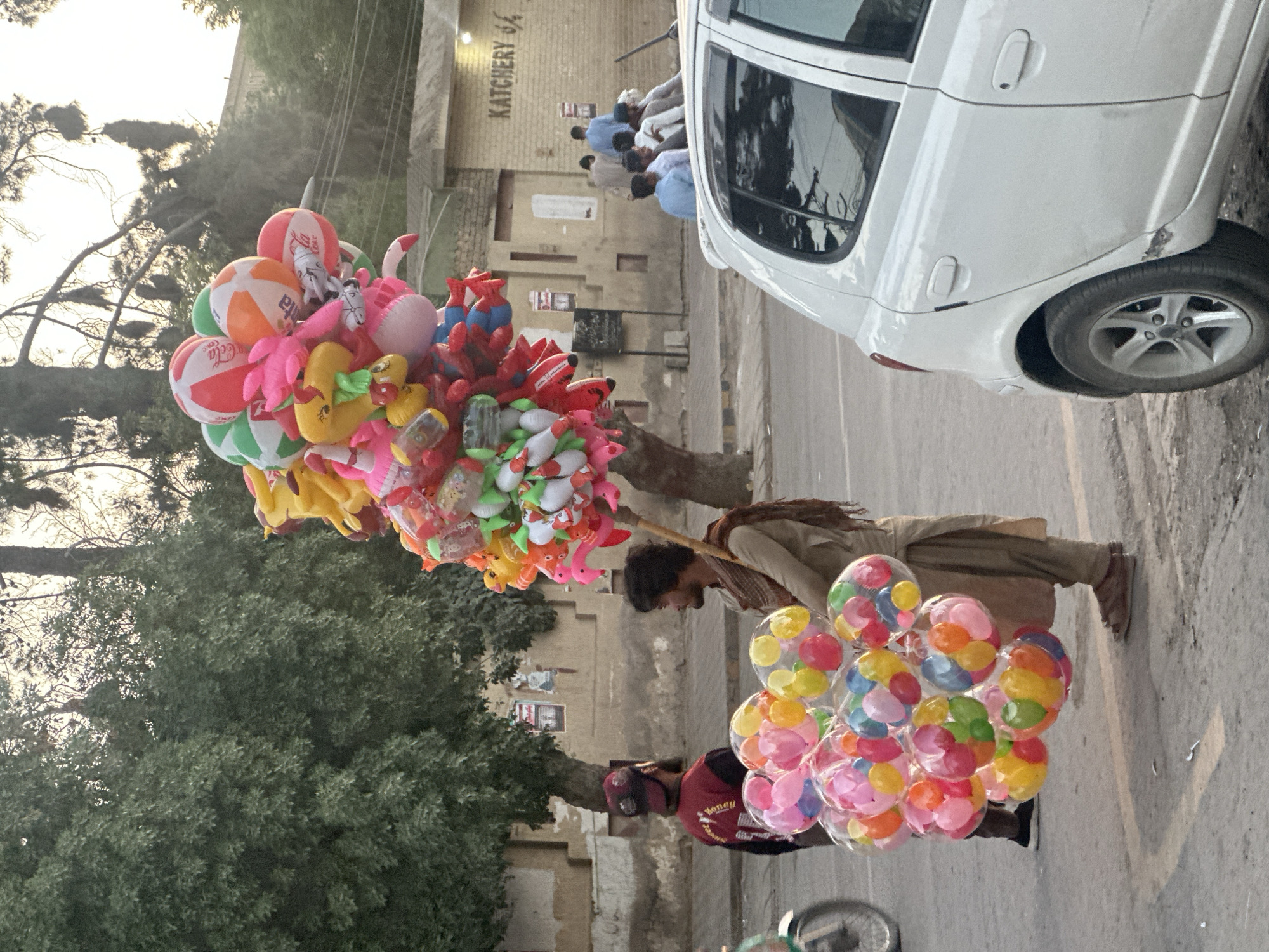 The image depicts two balloon vendors in Quetta City Tehsil, Pakistan. One vendor wears a tan outfit and carries a collection of inflatable toys and beach balls on a stick over their shoulder. The second vendor wears a red shirt and carries a large bunch of colorful balloons. They are standing on the street near a white car. In the background, a group of people are sitting outside a building with "Katchery 64" written on it. Trees and other buildings are visible in the distance.