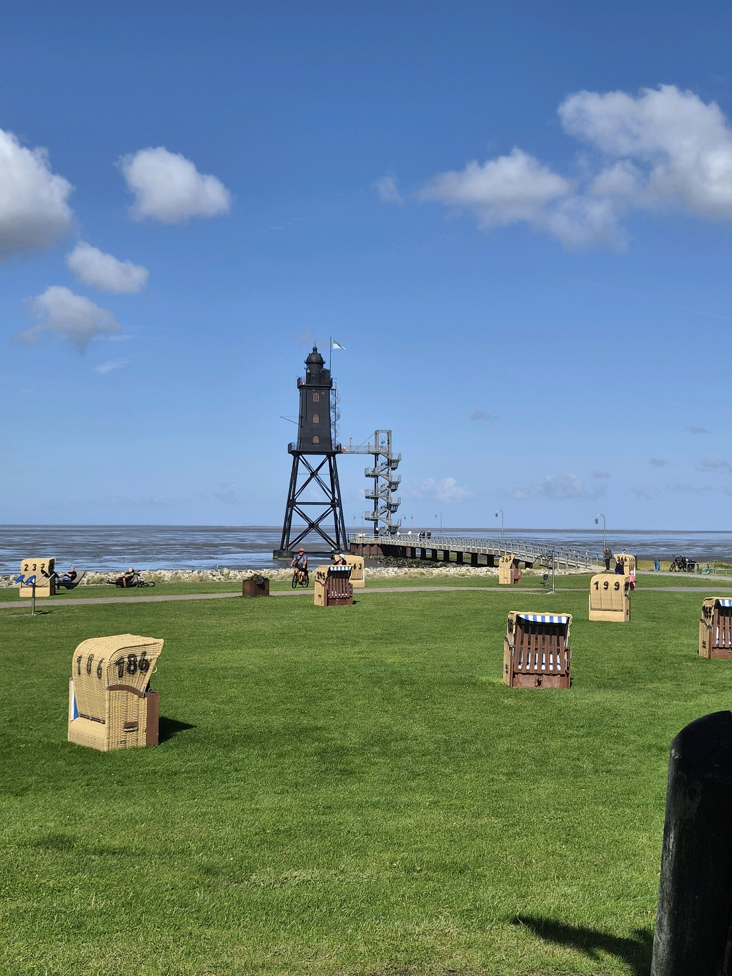 This image shows a wide shot of the coast at Dorum, Germany, on a clear day with scattered white clouds. In the background, a tall, black lighthouse with a white flag stands on a pier that extends into the calm, grey sea. The pier is accessed by a spiral staircase. In the foreground, a lush green lawn is dotted with numerous wicker beach chairs, some of which have numbers visible. Several people are visible on the lawn and along the path beside the beach chairs. One person is riding a bicycle, and others are relaxing in the chairs or lying on the grass. The overall scene depicts a peaceful coastal area with amenities for visitors.