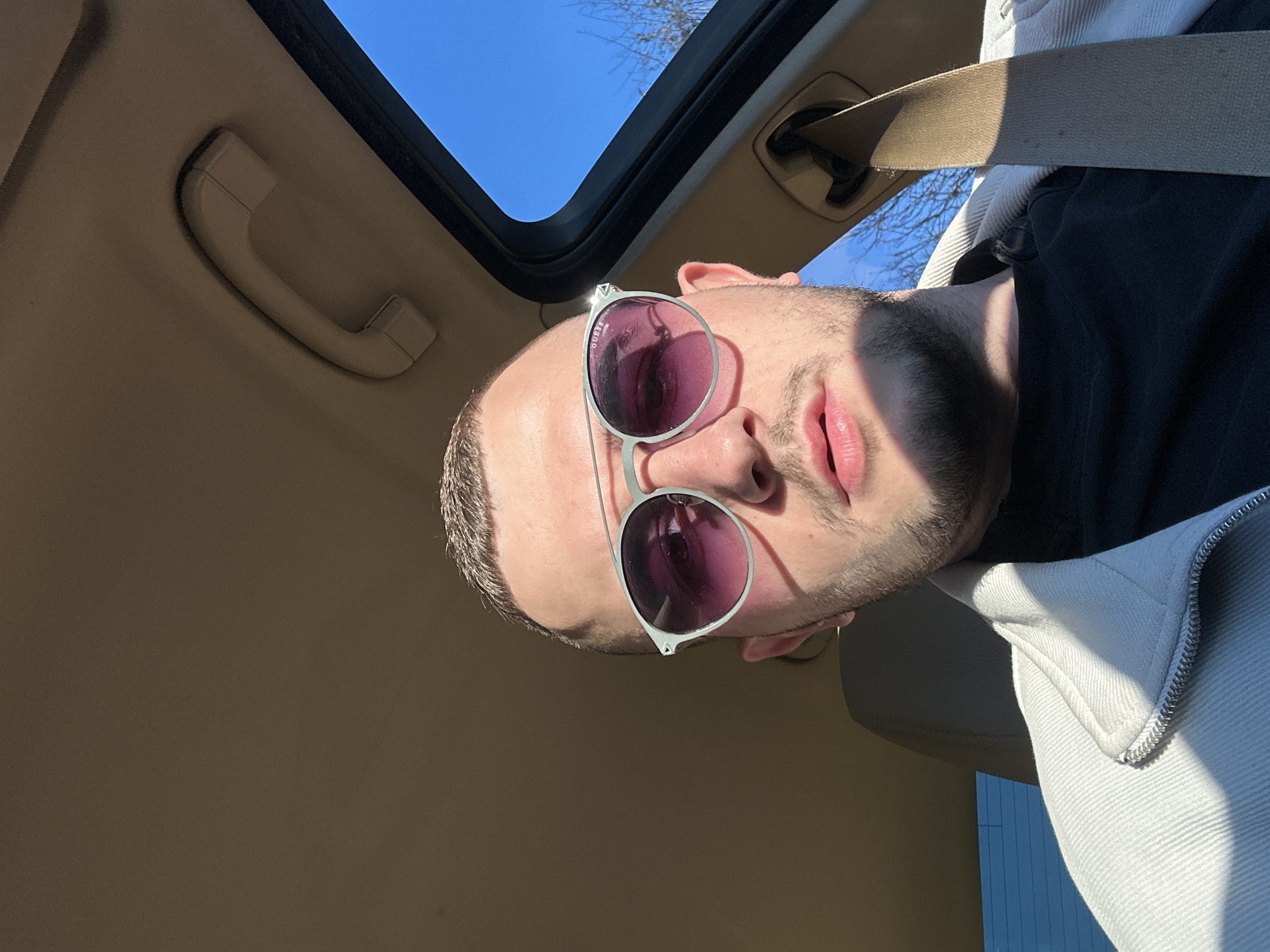 A low-angle, selfie-style shot depicts a person wearing sunglasses inside a car. The car's sunroof is open, revealing a clear blue sky. The person has short hair, a beard, and is wearing a black shirt and a light-colored jacket. The seatbelt is visible across their chest. The car's interior is beige. There is no indication that the location is Plovdiv, Bulgaria.