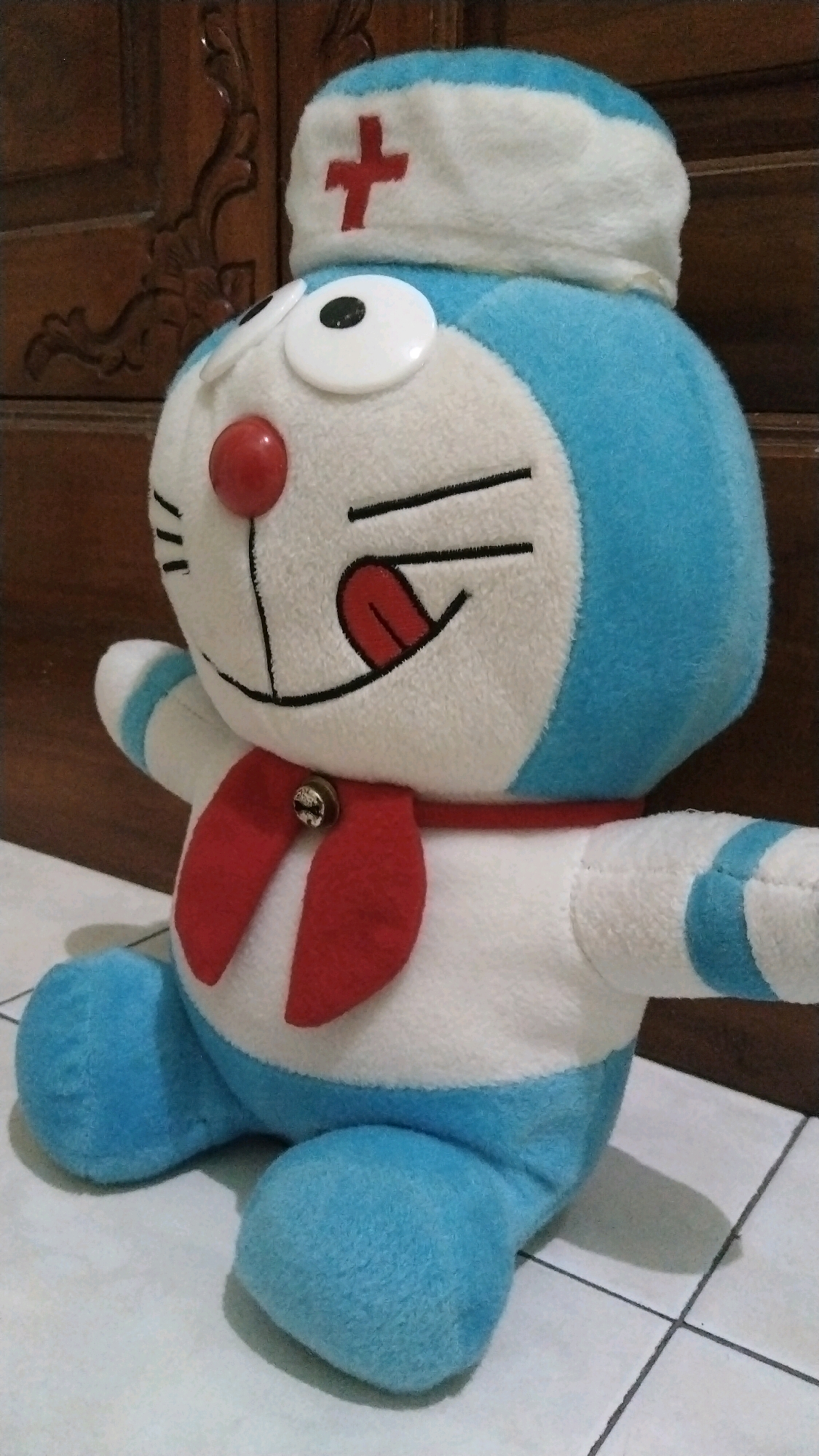 A plush toy resembling Doraemon, a character from a Japanese manga series, is positioned in a seated posture on a tiled floor. The toy is dressed as a nurse, wearing a white and blue nurse's cap with a red cross emblem and a red ribbon around its neck, adorned with a small bell. Its body is predominantly white and blue, with black stitching detailing its facial features. The background features a dark wooden cabinet with carved details, suggesting an indoor setting. The lighting appears to be natural, casting soft shadows. The image was taken in Simo, Indonesia.