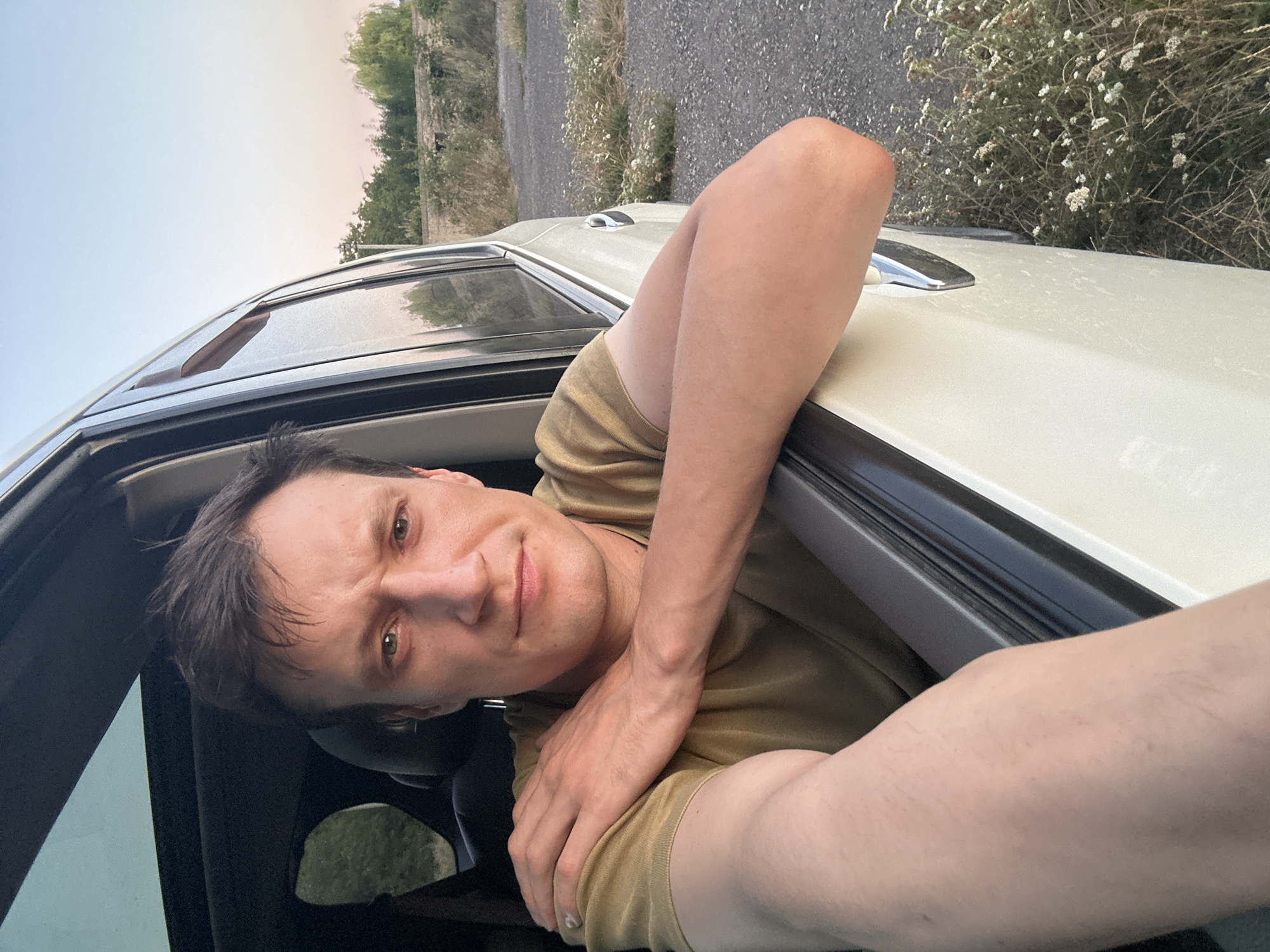 A man with fair skin and short, dark brown hair is looking out of the open window of a car. He is leaning his arms on the edge of the window frame, with his right arm resting on his left arm, which is bent at the elbow and resting on his shoulder. He is wearing a tan t-shirt. The car appears to be a light-colored SUV. The background shows a rural landscape with sparse vegetation and a pale blue sky. The image was taken in Makiv, Ukraine.