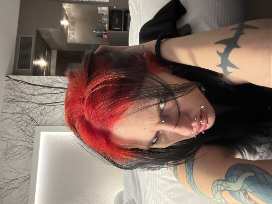 An individual with two-toned hair, bright red at the roots and black through the lengths, looks directly at the camera with a stern expression. Numerous silver-toned piercings are visible on their face, including the eyebrows, nose bridge, philtrum, and upper and lower lips. Dark eye makeup is present, with a subtle smudge beneath the left eye. Tattoos include a light blue coiled serpent or dragon on the upper left arm and a black barbed wire or thorny pattern on the upper right arm. The subject wears a dark, sleeveless top and has a black elastic band on their right wrist. They are positioned on a bed with white, textured bedding, with their left hand resting behind their head. The background reveals a modern hotel room in Plovdiv, Bulgaria, featuring a large, illuminated white headboard with a patterned surface. Above the headboard, the wall is decorated with a black and white graphic of bare tree branches. To the right, a mini-bar area contains bottles and glasses on a dark cabinet, situated below a rectangular air vent and two wall-mounted light fixtures. A dark wooden door is partially visible in the background.