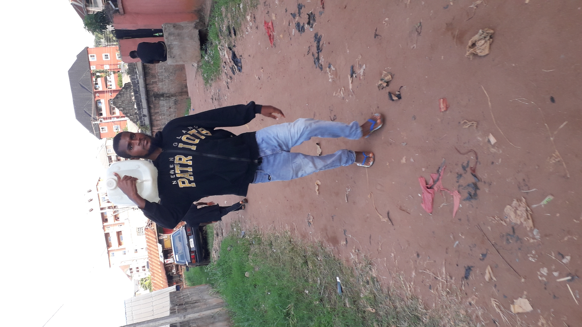 The image shows a man standing on a dirt road in Nkpor, Nigeria. The road is strewn with garbage. The man is holding a white plastic jerrycan. He is wearing a black jacket with yellow lettering, light blue jeans, and blue sandals. In the background, there is a multi-story building. A black car is seen parked on the side of the road. There is grass growing along the side of the road. Another person is visible on a ledge in the background.