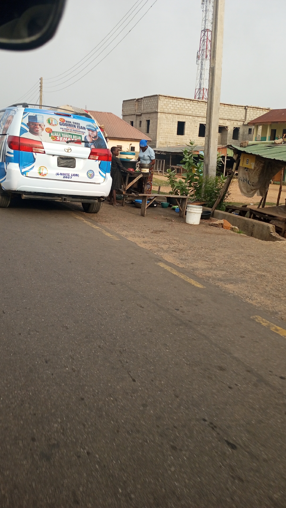 This image presents a street scene in Adankolo, Nigeria, captured during daytime under a bright, hazy sky. The foreground features a paved road with a yellow dividing line.
Dominating the left side is the rear of a white minivan or SUV, heavily decorated with political campaign livery. Prominently displayed are portraits of two men and accompanying text. The visible text includes "HON. CHIEF GODWIN ISAH" (with smaller, less distinct text possibly indicating "FOR HOUSE OF REPRESENTATIVE") and "ALHAJI YAHAYA BELLO FOR SENATE." Below these, the branding "(G-WHITE LION) 2027" is visible, suggesting a campaign for an election in that year.
To the right of the van, two people are present next to a wooden street stall. An adult, wearing a light blue head covering and top, appears to be preparing or serving something at the stall, with bowls and other items visible. A child stands beside the adult, observing the activity.
The background shows a mix of residential and commercial structures. An unfinished concrete building with multiple window and door openings stands prominently, alongside a building with a brown roof. Smaller, rustic market stalls line the side of the road. A tall communications tower or cell mast is visible in the distance, and power lines crisscross the sky. The overall scene depicts a typical bustling roadside environment.