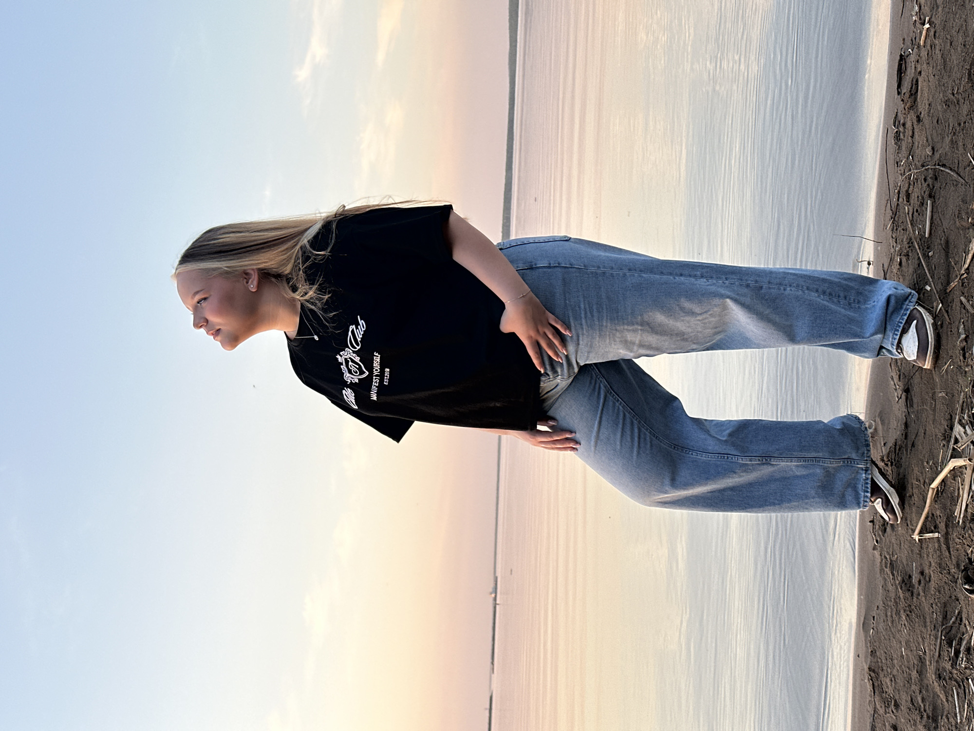 A full shot of a blonde woman in a black t-shirt and blue jeans standing on a beach. She is looking left, away from the camera. Behind her is a body of water with a light sky above. The beach is made of dark sand and some small pieces of debris. The image was taken near the city of Chaikovsky in Russia.