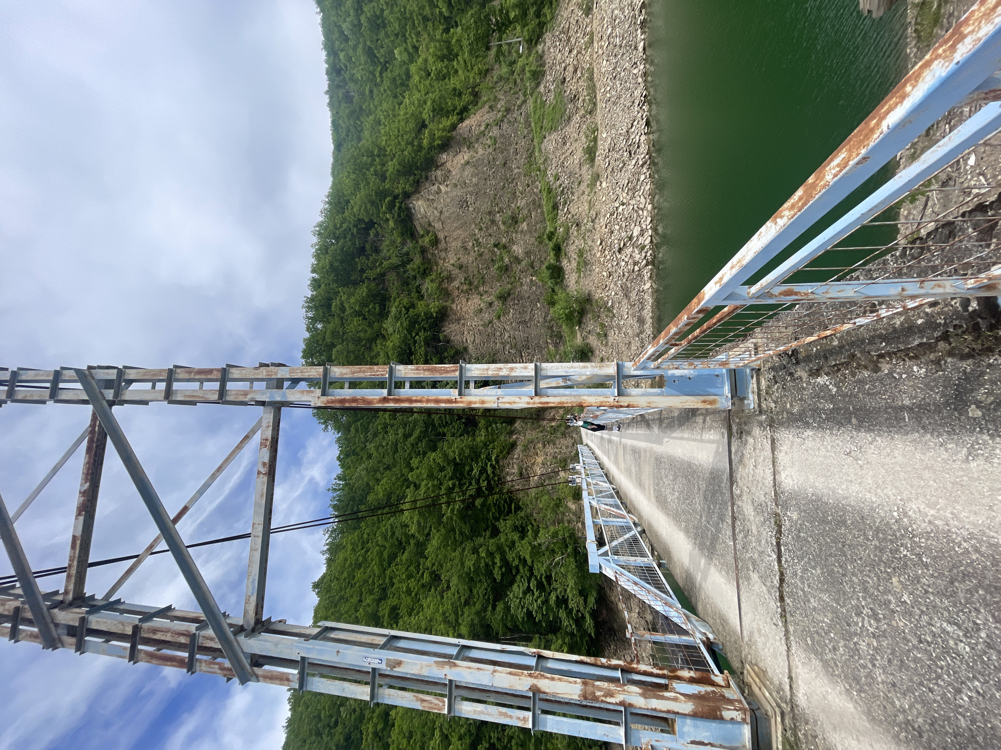 A vertical shot shows a bridge in Gračac, Bosnia and Herzegovina. It features a worn, blue and rusted steel structure with a concrete walkway. Lush green vegetation covers the adjacent hillside on one side, while the other side reveals a reservoir or lake with green-tinted water and a rocky shoreline. The sky is visible between the bridge structure, displaying scattered clouds.