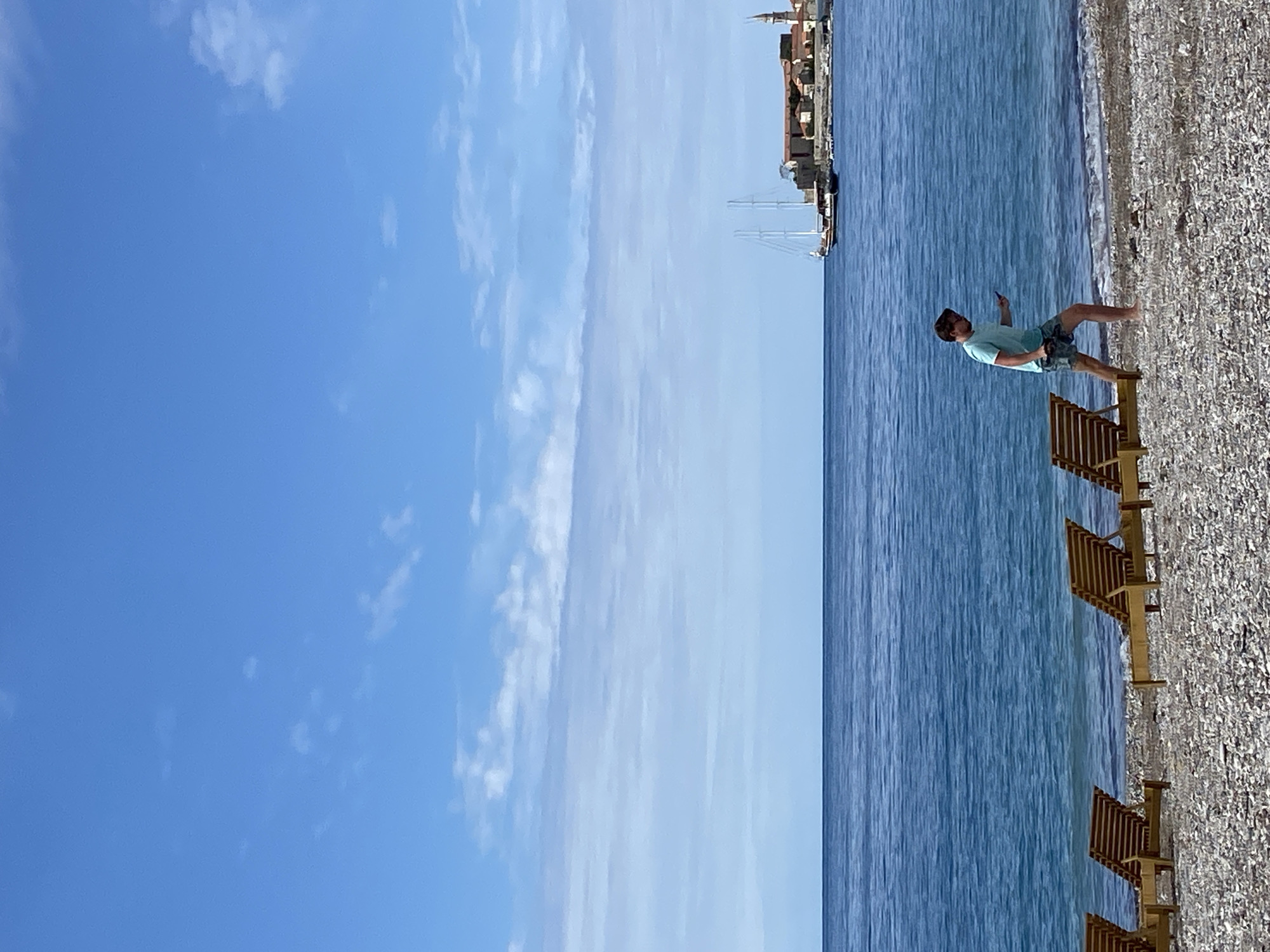A man stands on a beach in Budva, Montenegro. The man is wearing a light blue t-shirt and patterned shorts. He is barefoot and appears to be holding a phone. The beach consists of small stones. Several beach chairs are lined up on the sand near the water's edge. The sea is calm and blue. In the distance, there are buildings and a large sailboat. The sky is blue with some wispy white clouds.