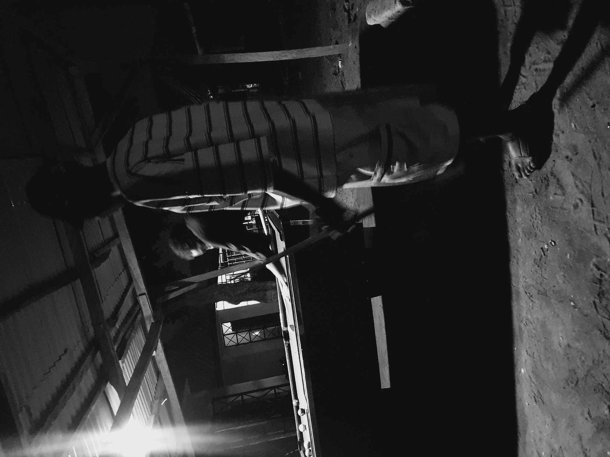A black and white photograph shows a man holding a long pole in Lokoja, Nigeria. He stands on a dirt ground next to what appears to be a building, possibly a school or apartment complex, viewed at night, in a dimly lit area. The man is positioned horizontally. The camera angle points upwards toward him. The man is wearing sandals. His shadow is cast behind him.