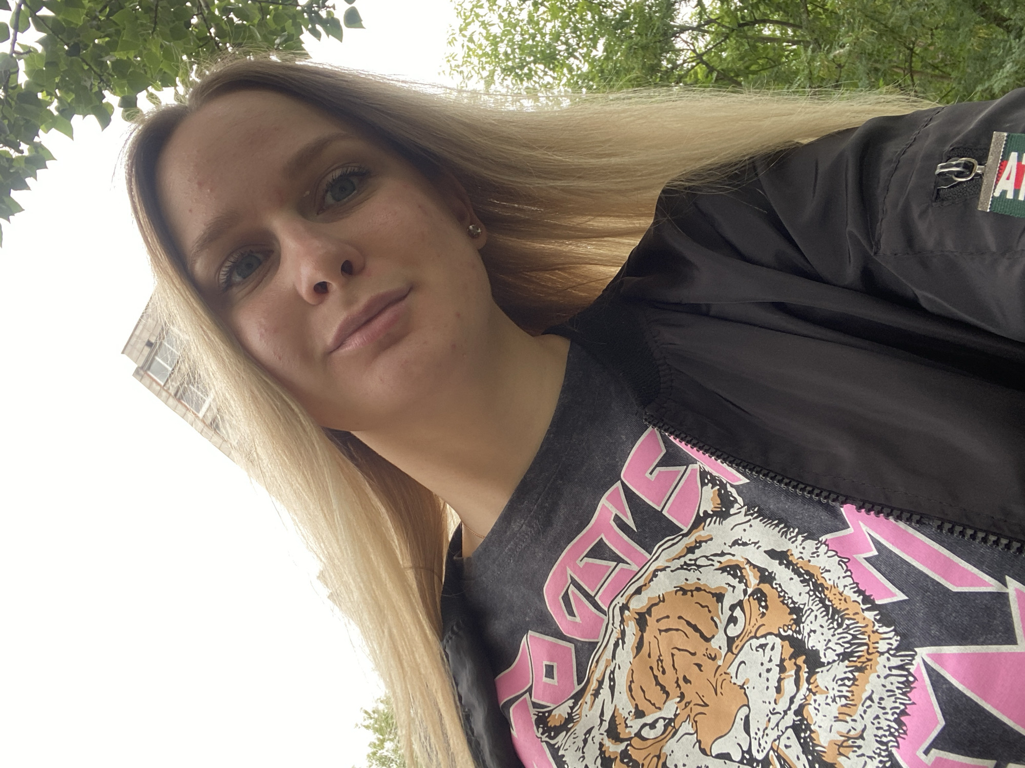 A young woman with long blonde hair and blue eyes is shown from the chest up, tilting her head towards the camera. She is wearing a black bomber jacket over a dark grey t-shirt with a graphic print of a tiger's face and pink lettering. The background consists of greenery and a building. The lighting is overcast.