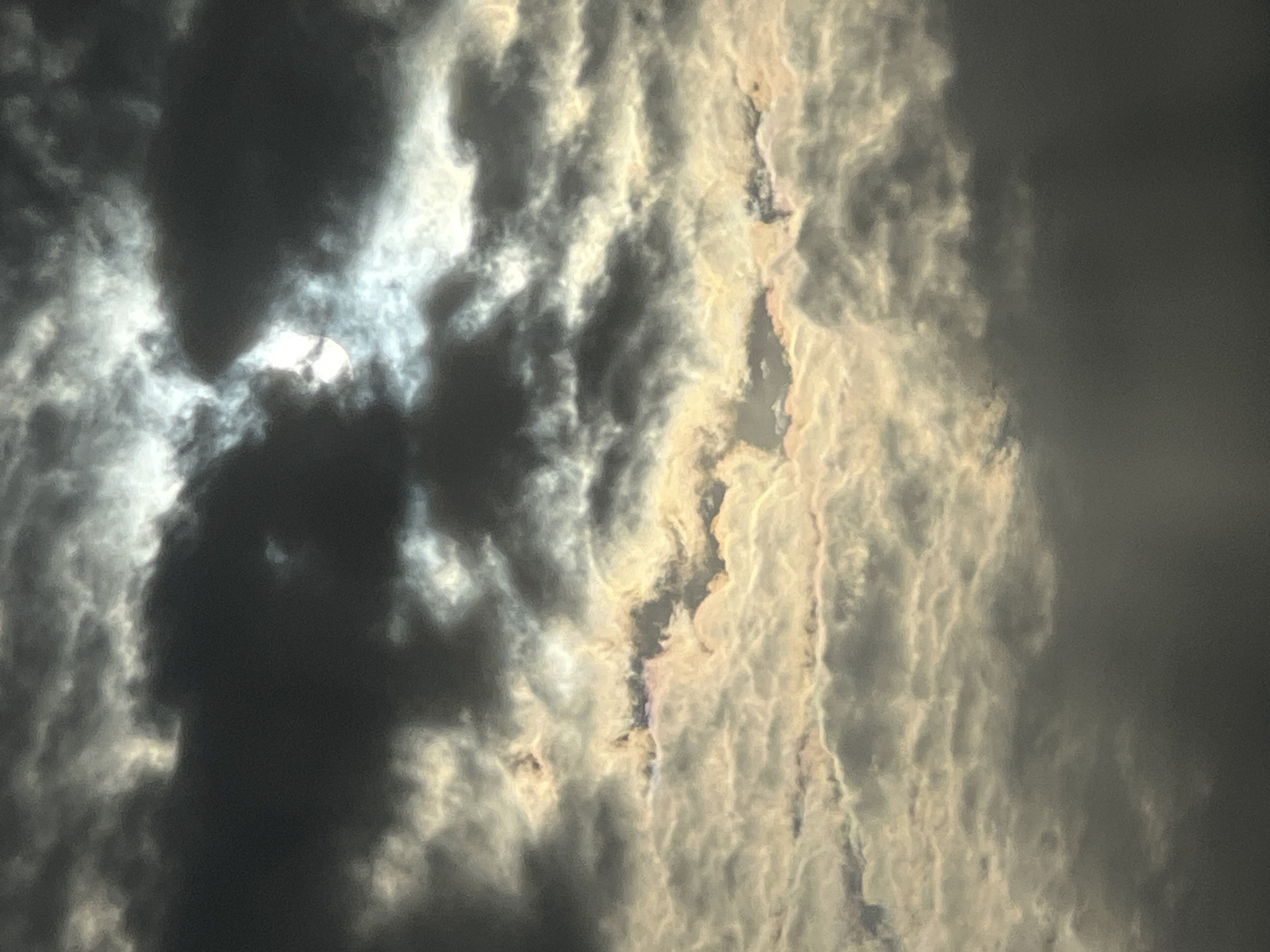 The image depicts a sky filled with clouds. Some clouds are dark and dense, obscuring the sun which is visible as a bright, obscured light source. Other clouds are lighter in color, reflecting the sunlight. There is no evidence of Krasnodar, Russia or any other location.
