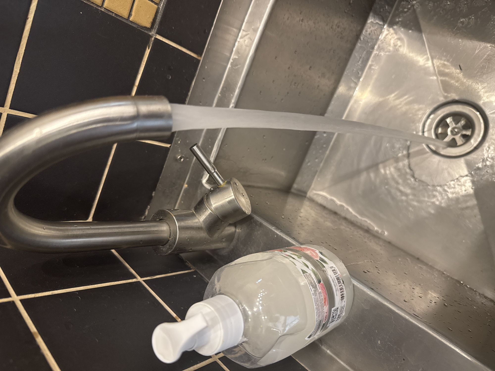 The image depicts a stainless steel sink with a curved chrome faucet dispensing water directly into the drain. A bottle of hand soap with a pump dispenser rests near the base of the faucet on the tiled surface adjacent to the sink. The tiles appear to be black with lighter-colored grout. The drain is a standard circular design with a star-shaped strainer. There is nothing in the image to indicate it is in the city of Yerevan, Armenia.