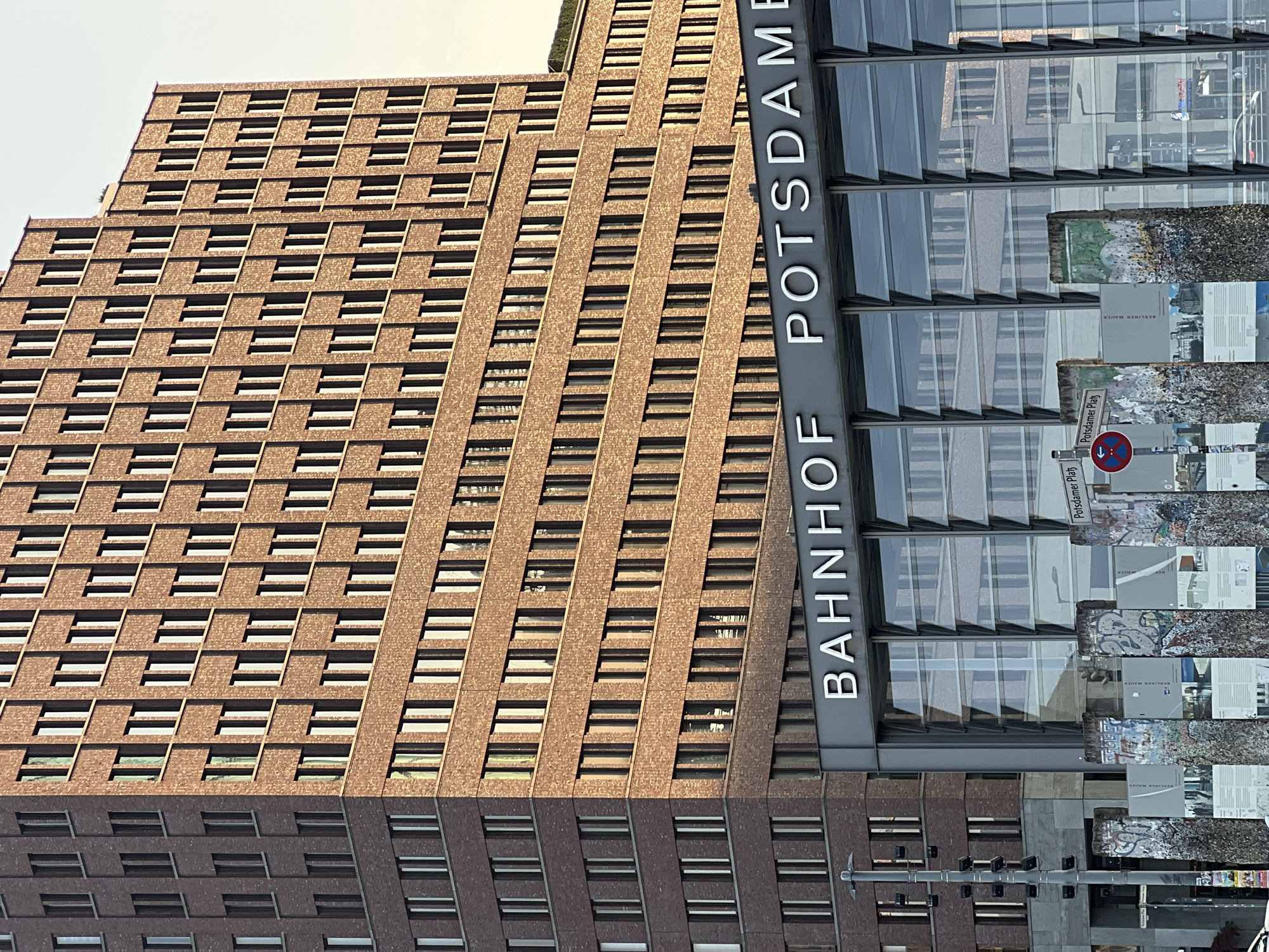 The image captures a section of Potsdamer Platz in Berlin, Germany, a location notable for its post-reunification architecture and historical significance. Dominating the scene is a tall, modern brick building with numerous evenly spaced windows, reflecting the warm glow of sunlight. Below this building, a contemporary structure with a glass facade displays the large white lettering "BAHNHOF POTSDAME," indicating a train station. In the foreground, remnants of the Berlin Wall, adorned with colorful graffiti and informational plaques, stand as a stark reminder of the city's divided past. The sky above is a pale, clear hue, suggesting a pleasant day. No people are visible in the frame.