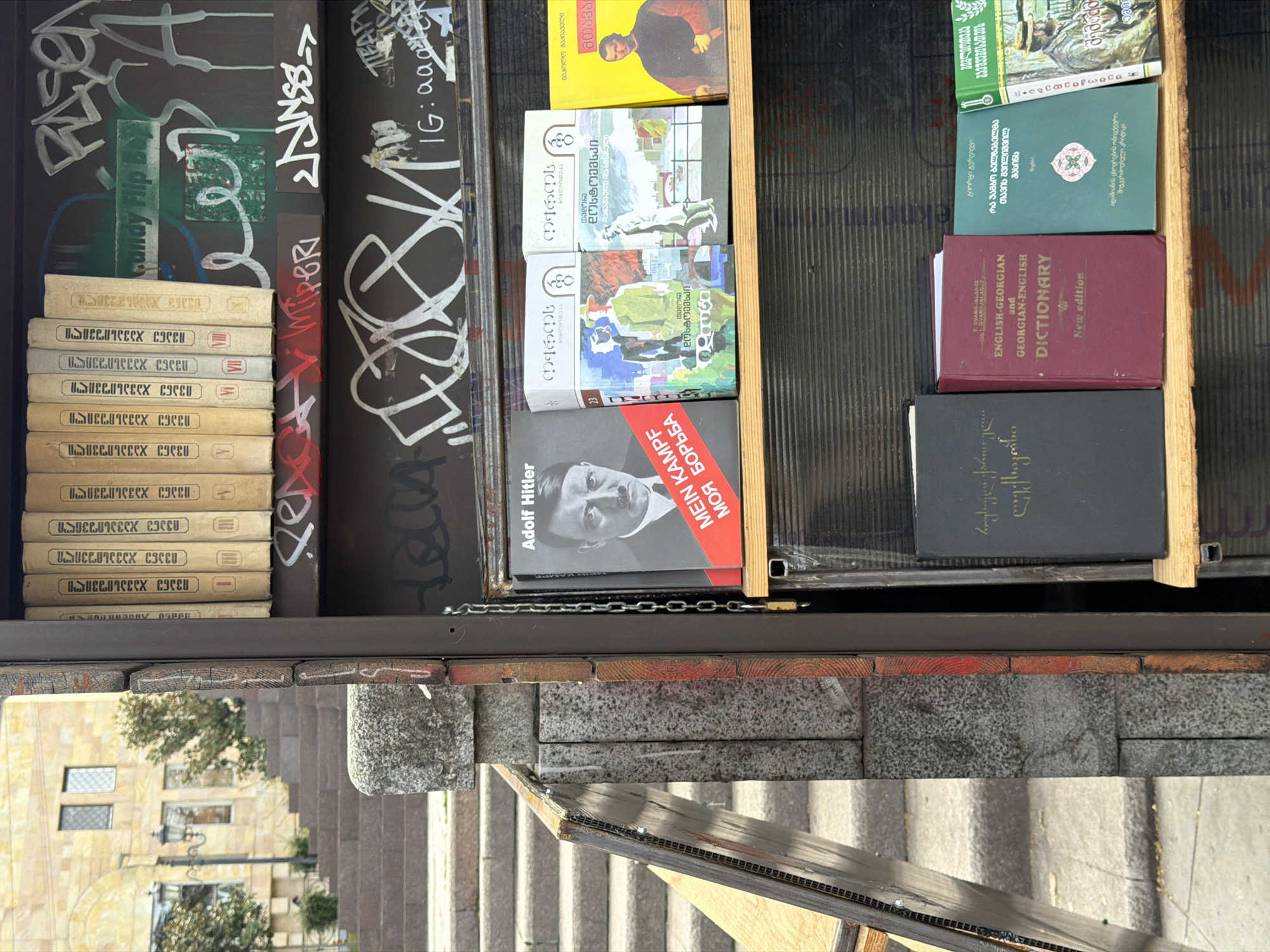 The image shows a small, outdoor bookshelf in Tbilisi, Georgia, located on a set of stairs. The shelves are filled with a diverse collection of books, ranging from classic literature to language dictionaries, including a copy of Adolf Hitler's "Mein Kampf." Graffiti covers the sides of the shelf, adding a layer of urban texture to the scene. The surrounding environment includes stone steps and visible parts of buildings and trees, characteristic of Tbilisi's urban landscape.