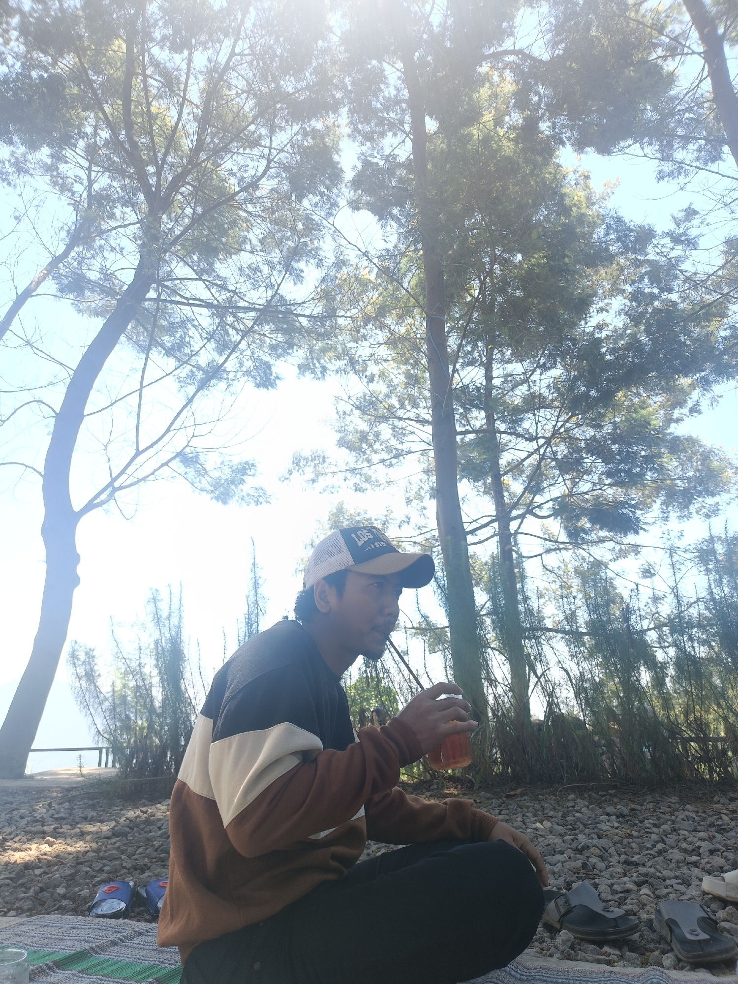 In a natural outdoor setting, likely the city of Kopeng, Indonesia, a person is seated on a striped blanket on a ground covered with small rocks. The person is wearing a long-sleeved shirt with a color-blocked design of brown, white, and dark blue, along with dark pants and a baseball cap. They are holding a clear plastic cup filled with a reddish-brown beverage, possibly tea, and appear to be drinking it. Surrounding the seated figure are various trees, with sunlight filtering through their branches and leaves, creating a bright, hazy atmosphere. In the background, there's a glimpse of a fence or railing, suggesting a boundary or overlook. Two pairs of footwear, one blue and white, the other gray, are visible on the ground near the blanket. The overall scene depicts a moment of relaxation amidst nature, possibly during a picnic or outdoor gathering.
