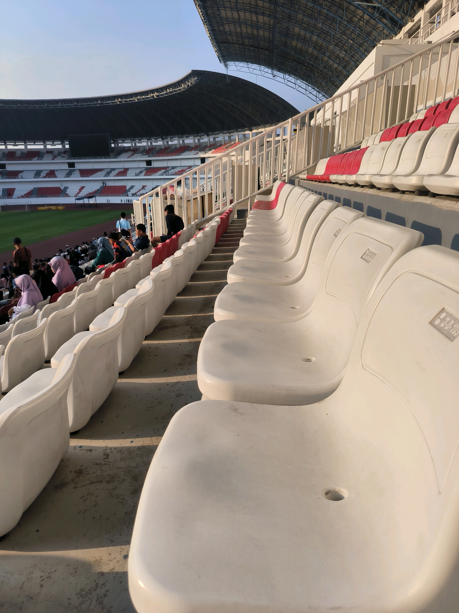 The image depicts an outdoor view of a stadium with tiered seating, likely located in Semarang, Indonesia. The seating area is comprised of rows of white plastic chairs, with some red seats visible in the background. A railing system runs along the side of the seating sections, separating them. The stadium has a large roof structure, partially visible at the top of the frame, supported by a metal framework. In the distance, a green sports field is visible. Several individuals are present in the stadium, some seated in the stands and others walking around. The lighting suggests it is daytime, possibly late afternoon due to the long shadows cast on the concrete floor and seating. Some of the white seats have visible markings or labels, such as "2-D-105". The environment appears to be clean and well-maintained.