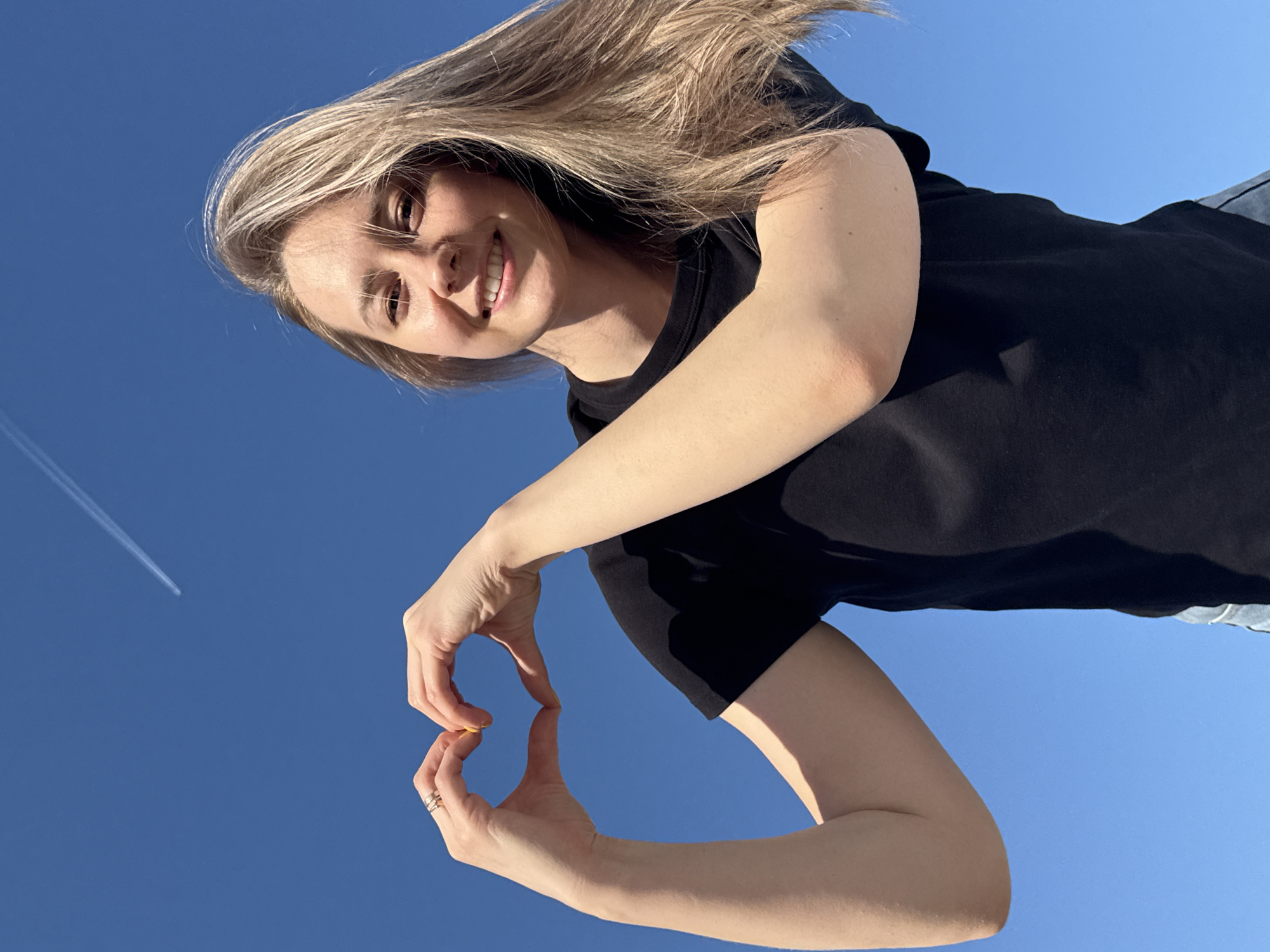A young woman is making a heart symbol with her hands against a clear blue sky. She is wearing a black t-shirt and has blonde hair. A jet trail is visible in the upper left corner of the frame. The specific location within the city of Chaikovsky, Russia cannot be determined from the image alone.
