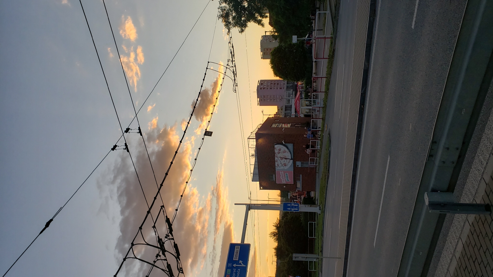 The image captures a street scene in Prešov, Slovakia, during sunset. The sky is a gradient of blue and orange, with fluffy clouds illuminated by the setting sun. Numerous overhead wires and poles, likely for public transport, crisscross the frame.
In the middle ground, a red brick building houses a "Redbox Pizza" establishment, indicated by a large advertisement on its facade. Several people are visible near this building, some standing and others sitting on a low wall. To the left of the pizza place, a blue street sign points towards "D1 E50" and "Sídliško III," suggesting a location within the city. Further to the right, two multi-story apartment buildings are visible. A Coca-Cola umbrella and a bus stop shelter are also present.
The foreground features a paved road with clear lane markings, a sidewalk, and a metal guardrail. The overall atmosphere is that of a typical urban evening, with the warm light of the sunset casting a soft glow on the surroundings.