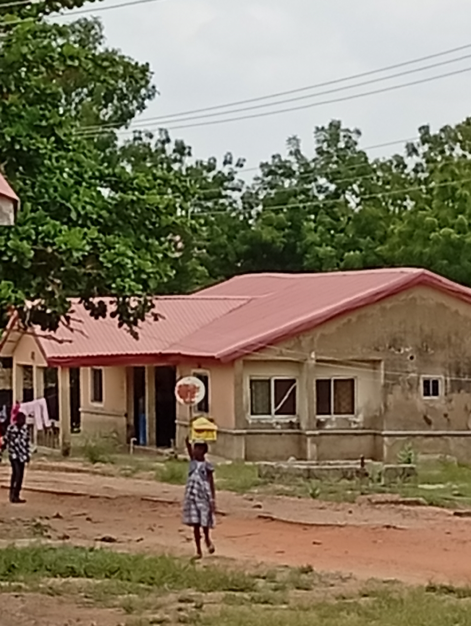 The image depicts a residential street in Adankolo, Nigeria. A single-story building with a red corrugated metal roof is in the background. The building's facade is unfinished, with visible cement. Two windows with white trim are on the right side. A person wearing a checkered shirt stands to the left. A young girl in a blue and white patterned dress is walking toward the camera, carrying a yellow container on her head. The ground is a mix of dirt and sparse grass. Trees surround the building and are visible in the background. Overhead wires are visible in the upper portion of the image. The sky is overcast.