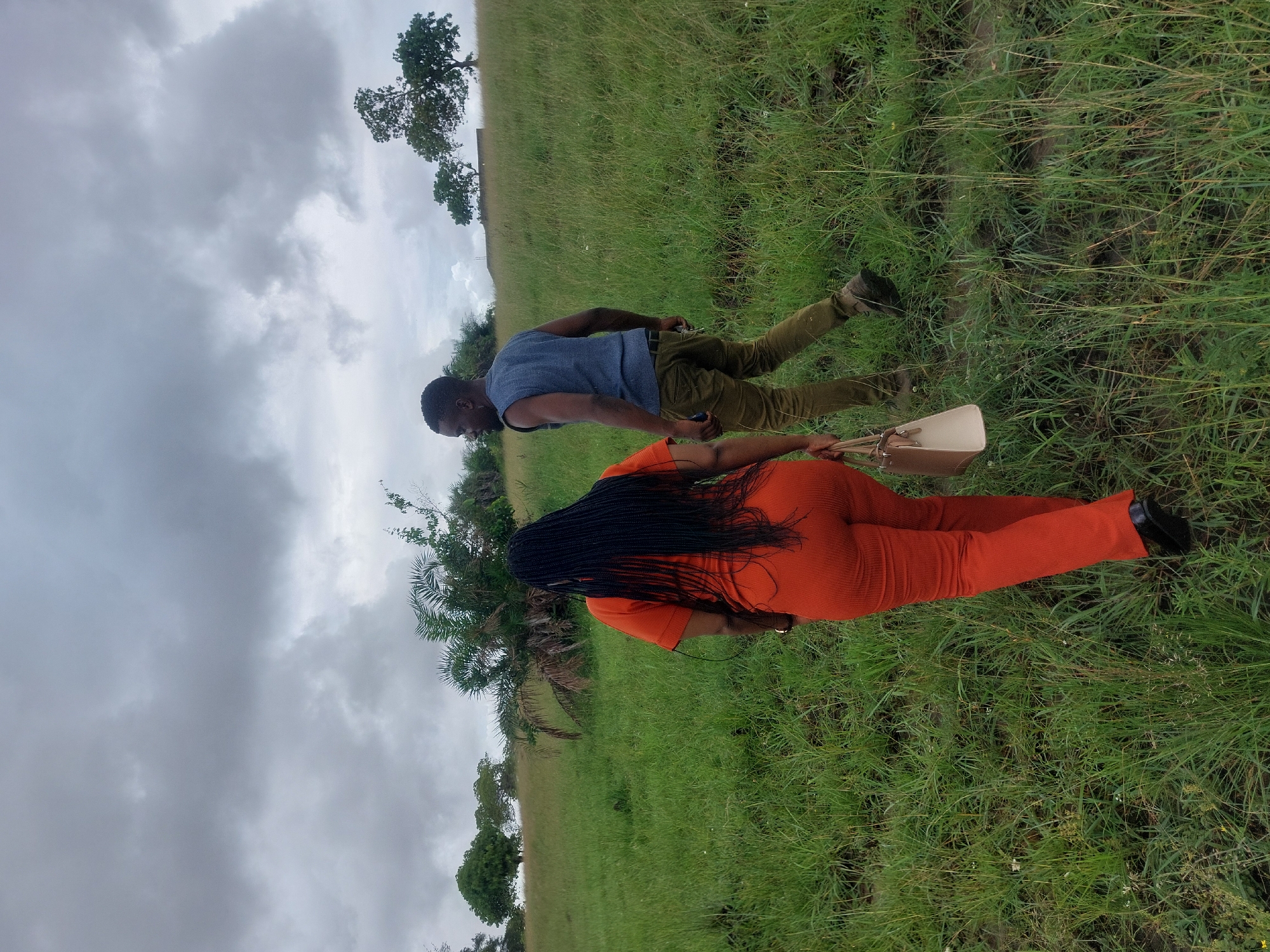 A man and a woman are walking in a grassy field on what appears to be a cloudy day. The man is wearing a blue vest and brown pants. The woman is wearing a bright orange outfit. She is carrying a light-colored purse. The field has scattered trees and some kind of structure in the background, but the overall atmosphere suggests a natural, open area. The picture was taken in Tanzania, but without further context, I can't pinpoint the exact location in Dar es-Salaam.
