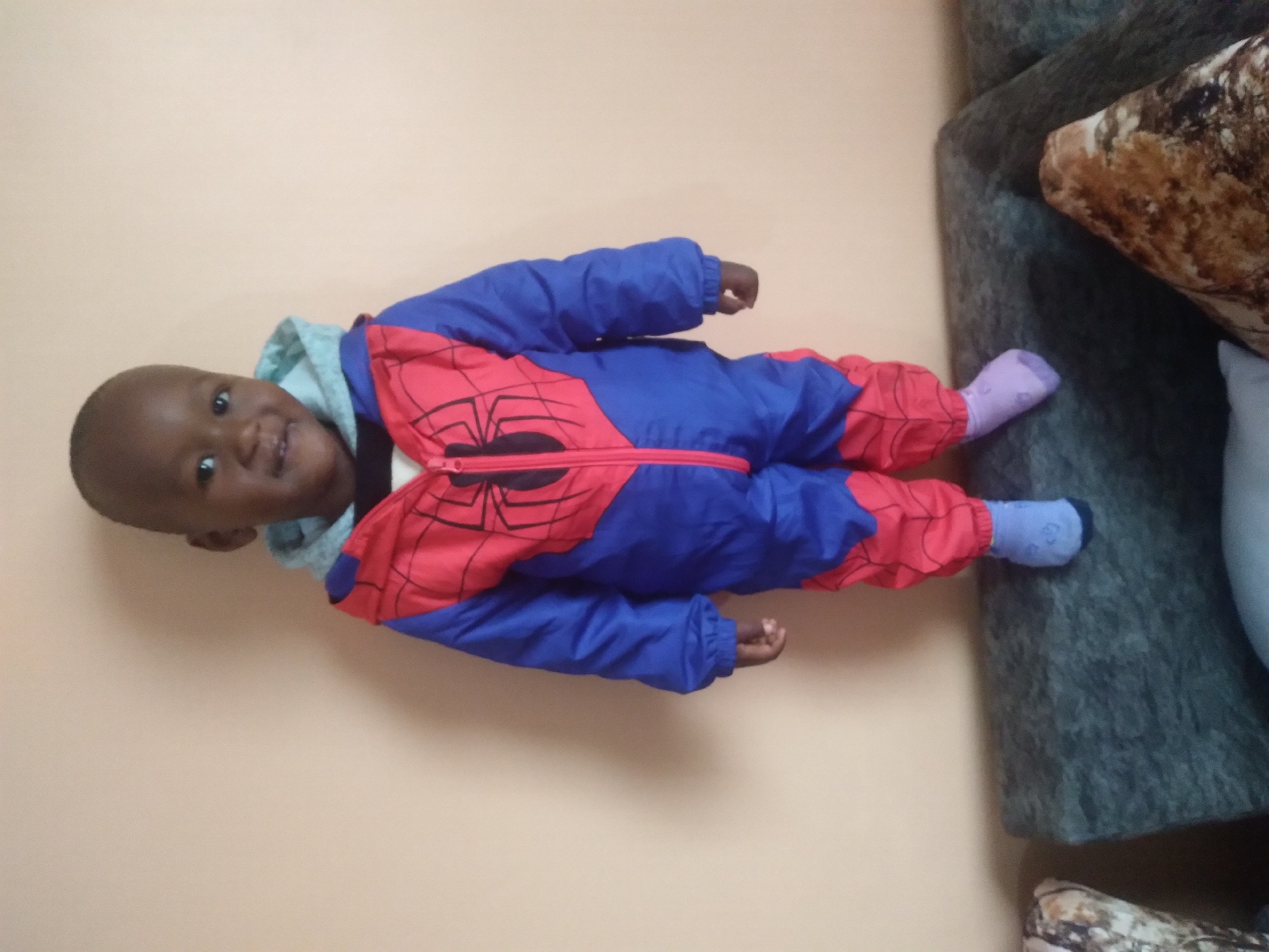 A young child, appearing to be around 2-3 years old, stands facing the camera in what seems to be a living room environment in Nyeri, Kenya. The child is wearing a blue and red Spider-Man themed jumpsuit with a grey hood. The child has dark skin and a shaved head, and is smiling. They are standing on the back of a dark grey sofa, with a plain, light orange wall as the background. To the left and right of the child, parts of patterned sofa cushions are visible. The lighting appears to be artificial, casting soft shadows.