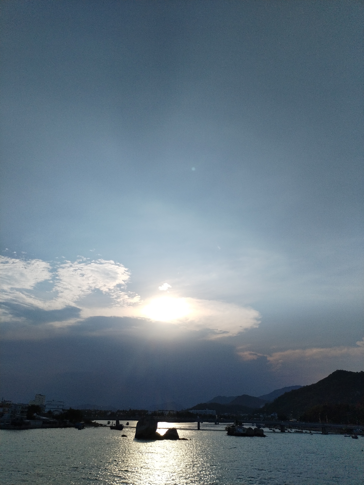 The image shows a body of water with scattered rock formations and a bridge, set against a landscape of mountains and urban development, likely in Nha Trang Ward, Vietnam. The sun is obscured by clouds, casting a bright glow and creating reflections on the water's surface. The sky transitions from a clear blue to darker, cloud-covered areas. Urban structures are visible on the left side of the frame, while mountains form the backdrop in the distance.
