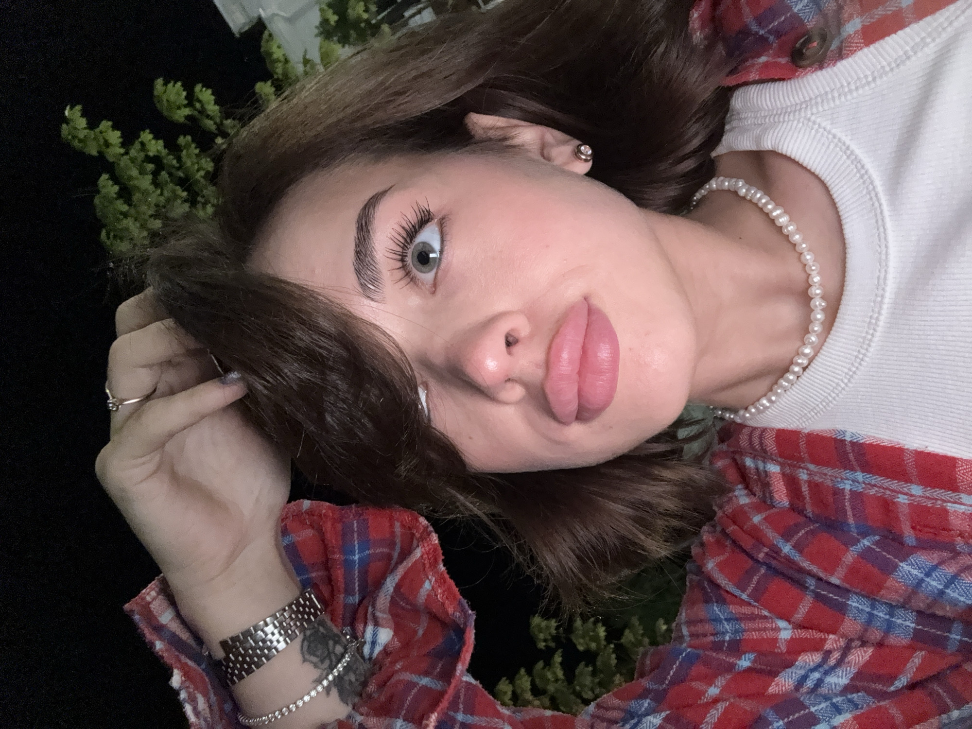 A young woman is seen in a tilted close-up shot. She wears a red and blue plaid shirt over a white top, and a pearl necklace. Her hair is brown and styled loosely around her face. She has light-colored eyes and full lips. Her left hand is visible, adorned with a ring, two bracelets, and a watch. The background is dark, with some green foliage visible in the upper left. There is no visible indication of Sarapul, Russia.