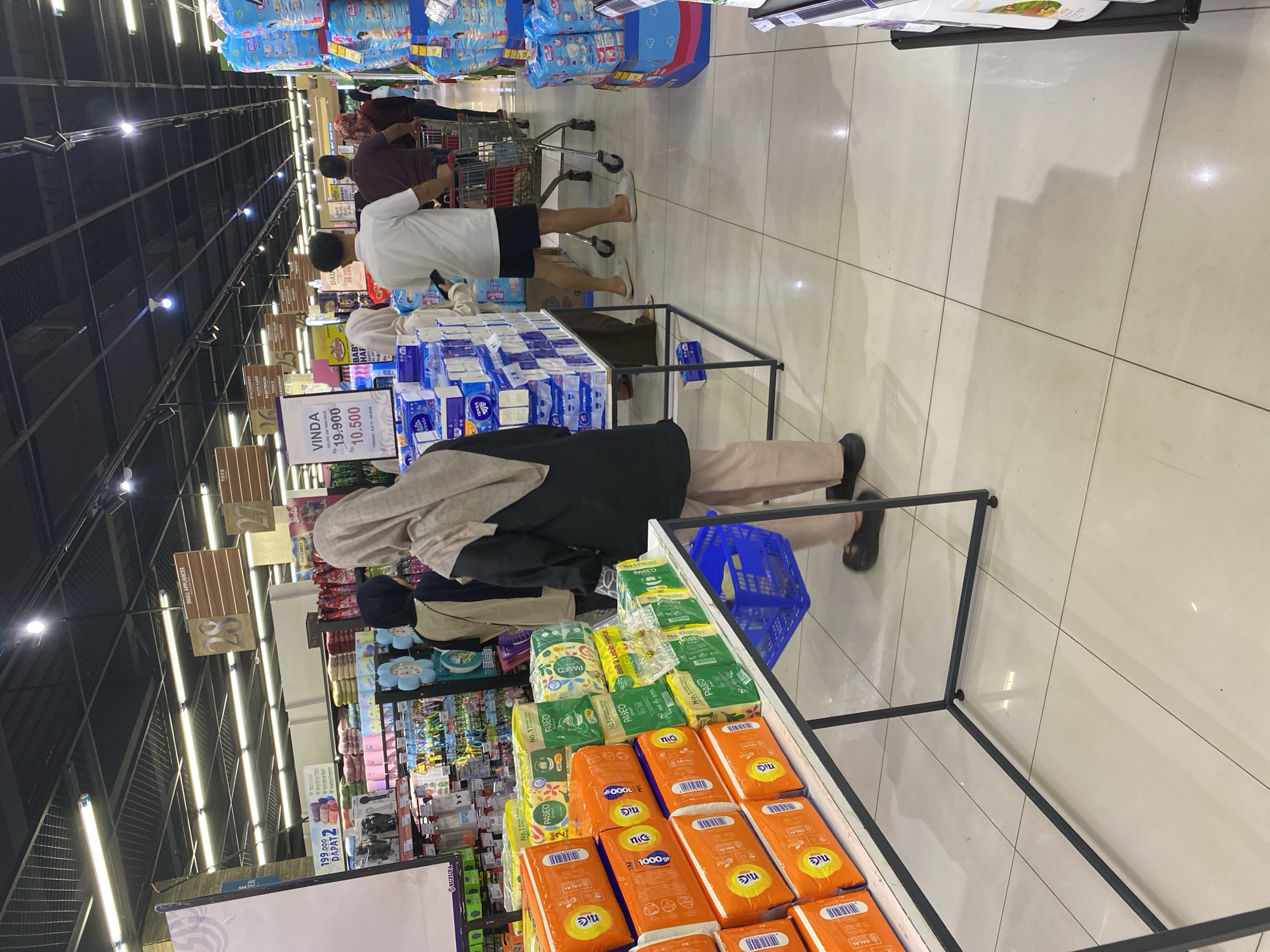 The image captures a wide-angle view inside a well-lit supermarket in Banda Aceh, Indonesia, with a focus on a display of tissue products and shoppers navigating the aisles. The ceiling features a modern grid of fluorescent lighting. In the foreground, on the left, a white metal shelf unit displays stacks of orange and green tissue paper packages, with prominent branding like "Nice" and "Paseo." A blue plastic shopping basket sits on the lower shelf of this unit.
Several individuals are present in the midground and background. A woman wearing a beige hijab and a dark jacket, along with light-colored trousers, is seen from behind, facing away from the camera and towards the tissue display. To her left, another woman, also wearing a hijab, is partially visible. Further back, a man in a white t-shirt and dark shorts pushes a shopping cart, followed by other shoppers.
Overhead signage indicates store sections, with wooden signs displaying numbers and categories like "SMALL APPLIANCES," "28," and "27." A prominent sign on a pink stand reads "VINDA" with pricing information for "DELUXE 250 THIN PACK" at Rp. 19,900 and a discounted price of Rp. 10,500, valid until 20-10-2021. The floor is tiled with a neutral color, and shadows from the ceiling lights and merchandise create a dynamic visual effect. To the right, stacks of blue and white packaged diapers are visible. The overall environment suggests a typical retail setting with customers engaged in shopping.