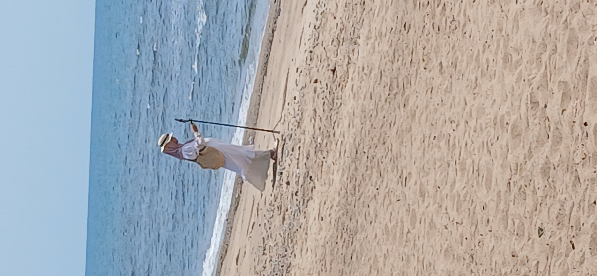 A woman wearing a white abaya, pink hijab, and a straw hat walks along the sandy shore of the sea. She carries a straw bag over her shoulder and uses a black walking stick for support. The sea is blue with gentle waves, and the sky is clear. The sand is light brown and textured with small pebbles and debris. The image captures a serene moment of a solitary figure enjoying the coastal environment. The location is identified as Banyuglugur, Indonesia.