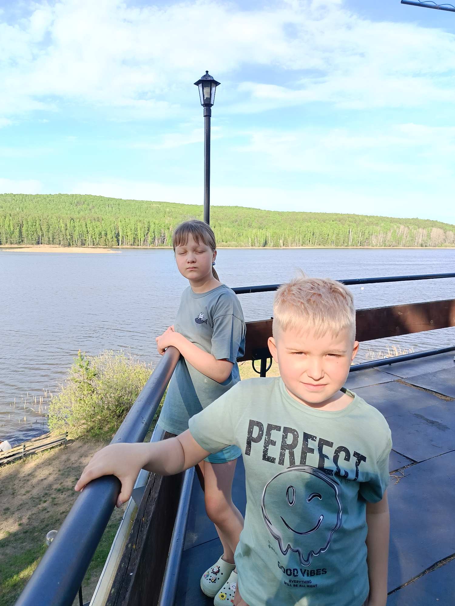 Two children are standing on a deck overlooking a body of water, presumably in the city of Chaykovsky, Russia. The boy in the front is fair-haired, wearing a green "PERFECT" t-shirt. The girl behind him has darker hair and is wearing a similar green shirt. They are both holding onto the railing of the deck. In the background is a lake or river with a forested shoreline. A lamppost is also visible. The weather appears sunny.