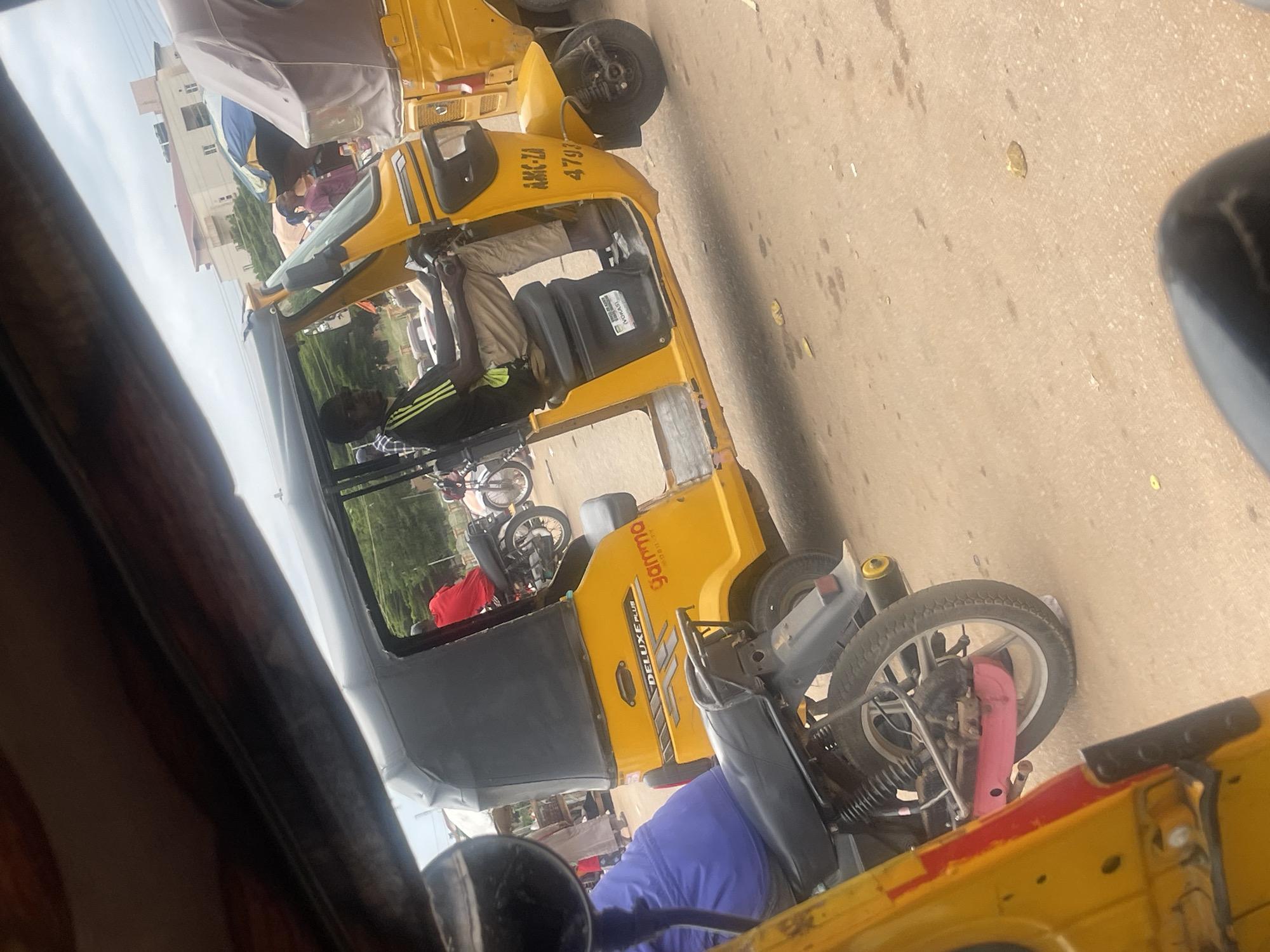 The image captures a side view of a yellow auto-rickshaw, likely taken from inside another vehicle. The auto-rickshaw is parked on a paved surface, possibly a road or street, in Galadimawa Village, Nigeria. Several people are visible within and around the auto-rickshaw, suggesting it is in use or waiting for passengers.
The subjects include a driver in tan pants and a black and green shirt, seated at the controls of the auto-rickshaw. Another person in a blue garment is partially visible behind the driver, possibly a passenger. In the background, several other auto-rickshaws and motorcycles are parked, with some people standing around them. A building with multiple stories is also visible in the distance, suggesting an urban or semi-urban setting.
The auto-rickshaw itself is bright yellow with black seating and some visible signage. The license plate reads "AKK-ZA 4793". There is also text on the side of the vehicle, including "DELUXE PLUS" and what appears to be "Ganno" in a stylized font.
The lighting suggests it is daytime, and the paved surface appears dry, indicating fair weather. The scene depicts a typical day in a bustling area where auto-rickshaws are a common mode of transportation. The overall impression is one of everyday activity and local commerce.