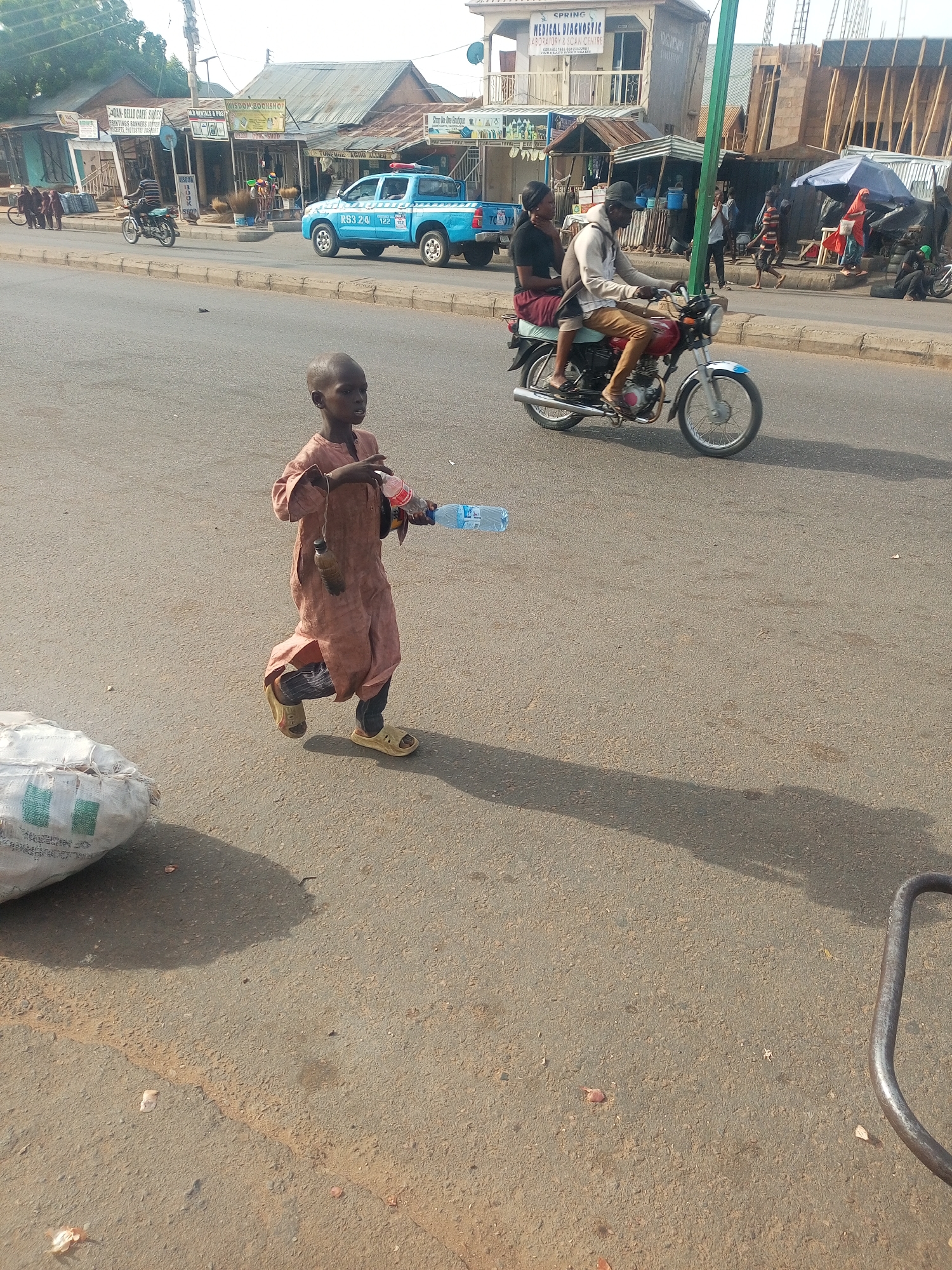 The image depicts a street scene in Tempure, Nigeria. A young child in a light brown robe and sandals is walking towards the camera, holding plastic water bottles. A motorcycle with two riders is traveling in the same direction as the child. A light blue pickup truck is parked on the side of the road. Buildings line the street, including a "Wisdom Bookshop," "Spring Medical Diagnostic" center, and other businesses with signs in English. Various people are walking and gathered in the background. A large bag and a metal object are in the lower portions of the image.