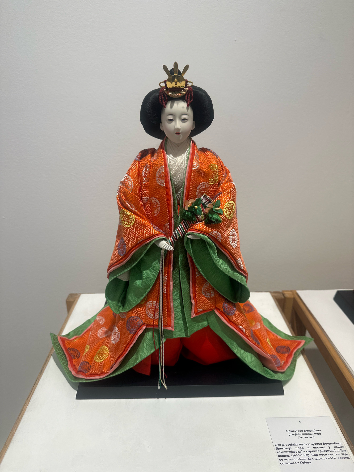 The image shows a Japanese Hina doll, likely an Empress, on display in the Museum of Yugoslav History (now the Museum of Yugoslavia) in Belgrade, Serbia. The doll is elaborately dressed in an orange kimono with green underlayers, adorned with intricate patterns. It wears a black wig and a golden crown. The doll is positioned on a black platform, which is placed on a white display surface. In the background, a white wall is visible. A descriptive plaque in Cyrillic script is placed in the lower right corner of the image, identifying the doll and providing some historical context.