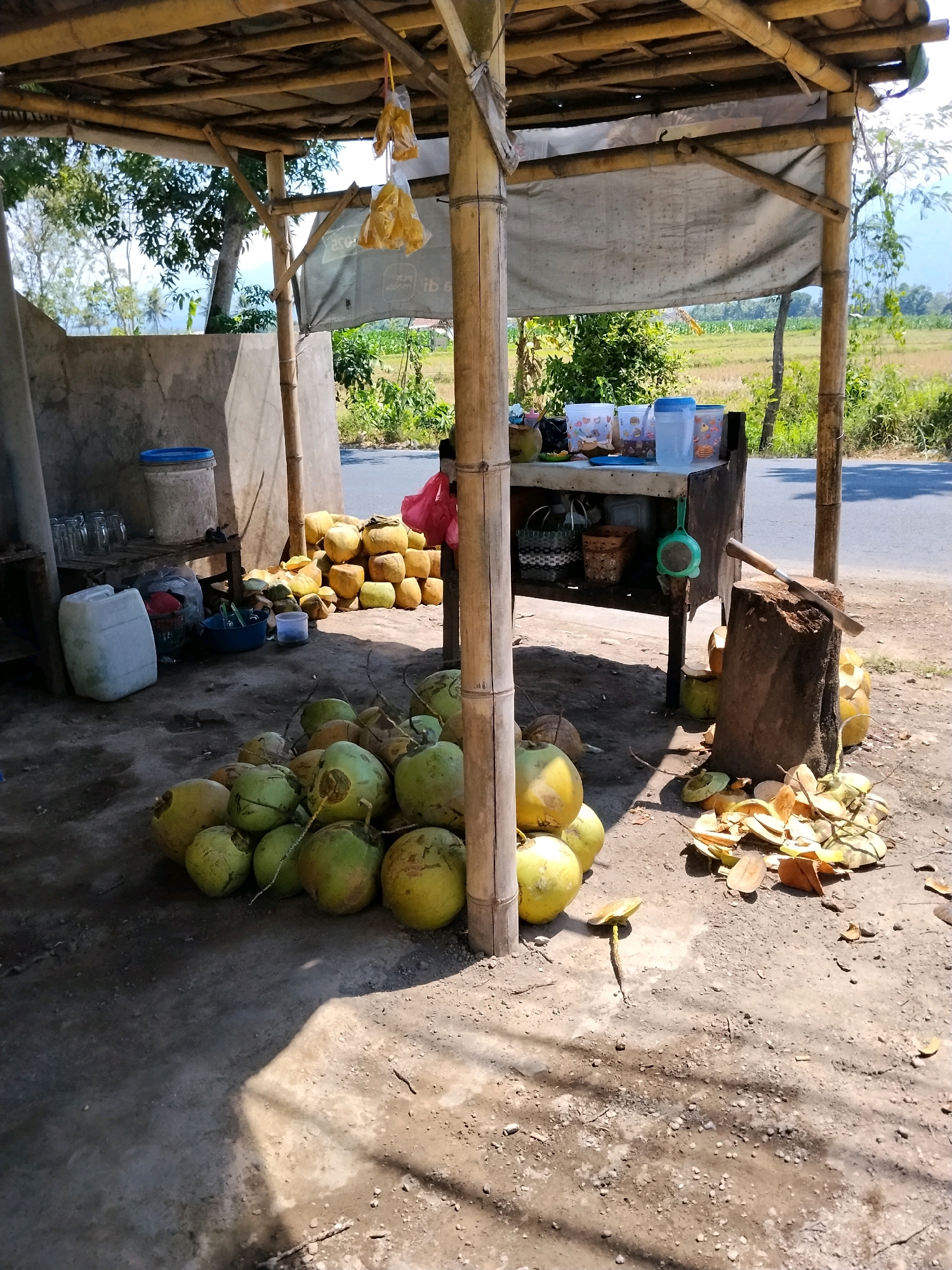 A scene in Kejawan, Indonesia, features a roadside stall constructed of bamboo and covered with a tarp. A pile of coconuts sits on the ground in the foreground, with some already opened and their husks scattered around a chopping block. A cleaver rests on the chopping block. The stall itself displays various containers, some holding what appear to be beverages or snacks, as well as jars and a large white plastic container. In the background, a dirt road runs alongside a field, with trees and vegetation completing the rural landscape. The overall impression is one of a small, functional business operating in an open-air environment.