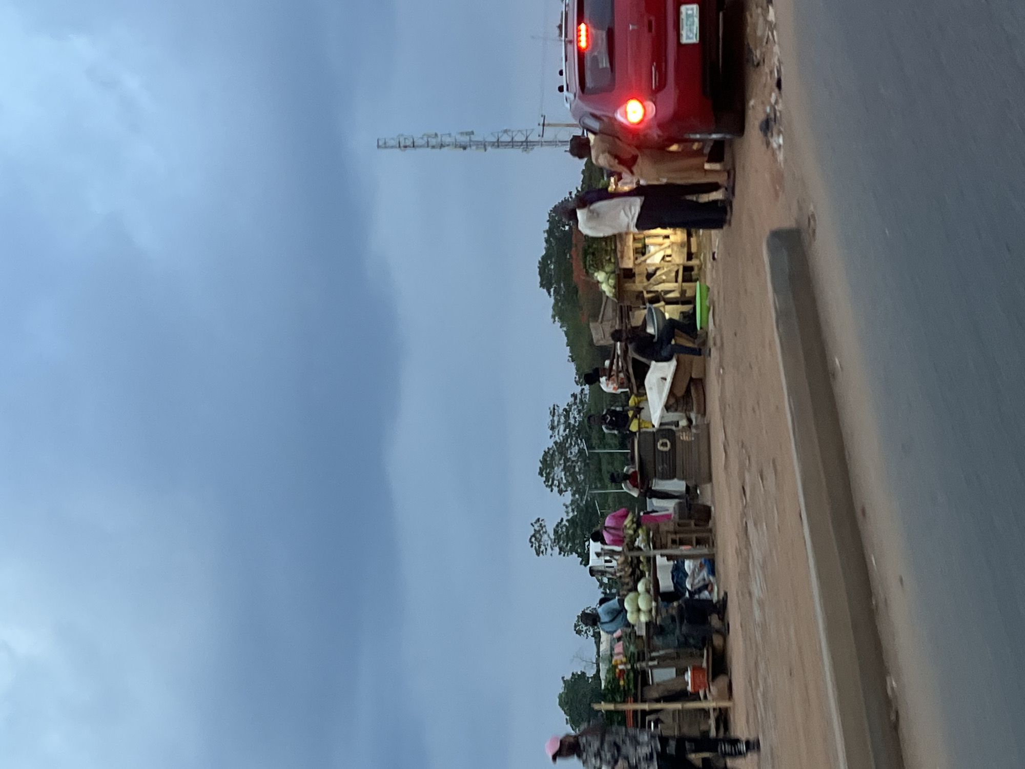 The image depicts a roadside scene in Galadimawa Village, Nigeria. A row of market stalls lines the edge of a road, with people gathered around them. The stalls appear to be selling produce, including melons and other vegetables. A red vehicle is parked on the road, its brake lights illuminated. In the background, there's a cell tower. The sky is overcast. The interaction suggests typical commercial activity. The environment suggests a semi-rural or peri-urban setting.