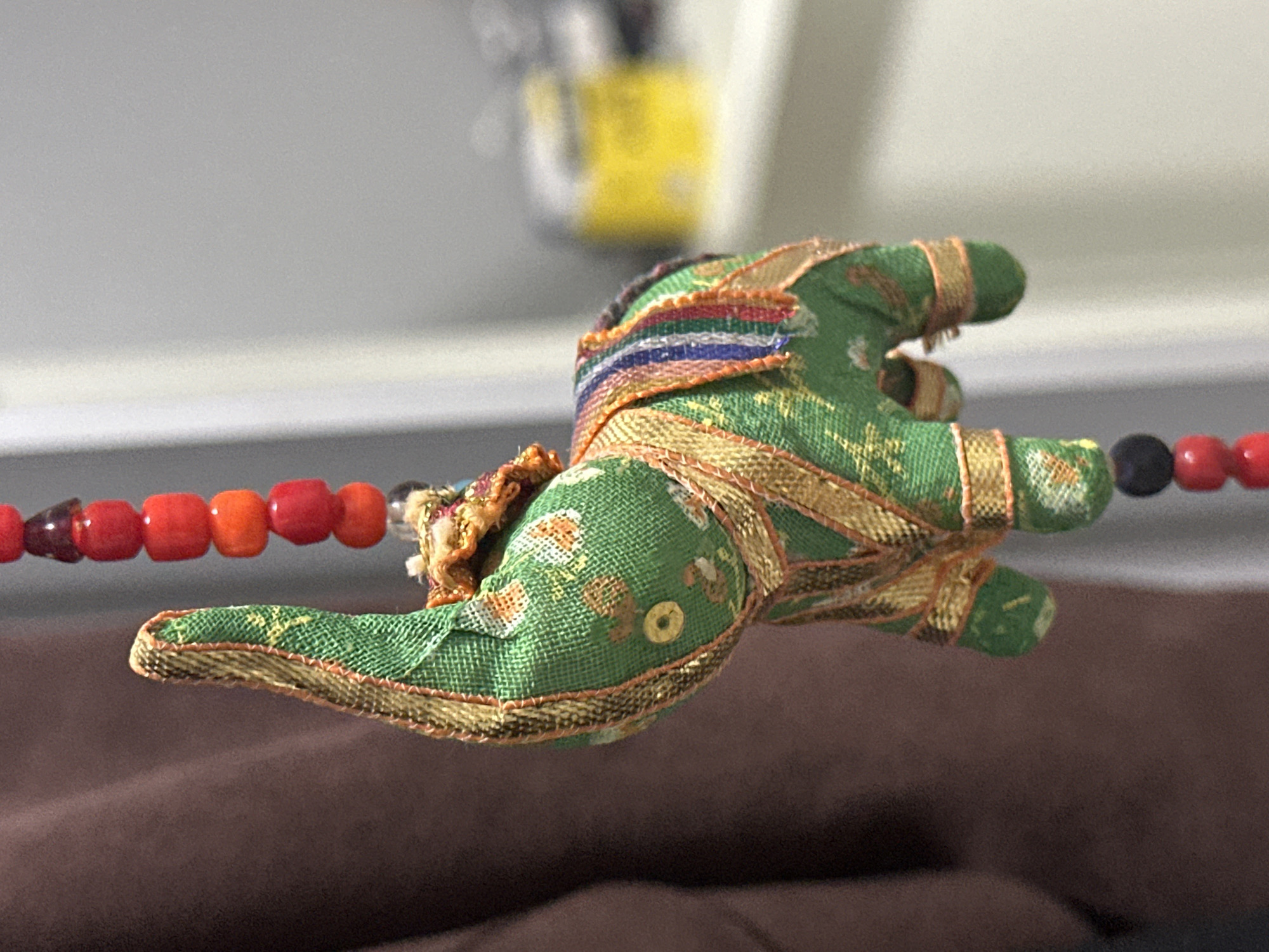 A close-up, shallow depth of field shot shows a green fabric elephant ornament hanging. The elephant is decorated with gold trim and a colorful striped fabric saddle. It hangs from a string of beads, with red, orange, and dark beads visible. The background is blurred, showing a brown textured surface on the left and a white vertical surface on the right, with a yellow rectangular object out of focus in the middle distance. There are no people or discernible actions in the image.