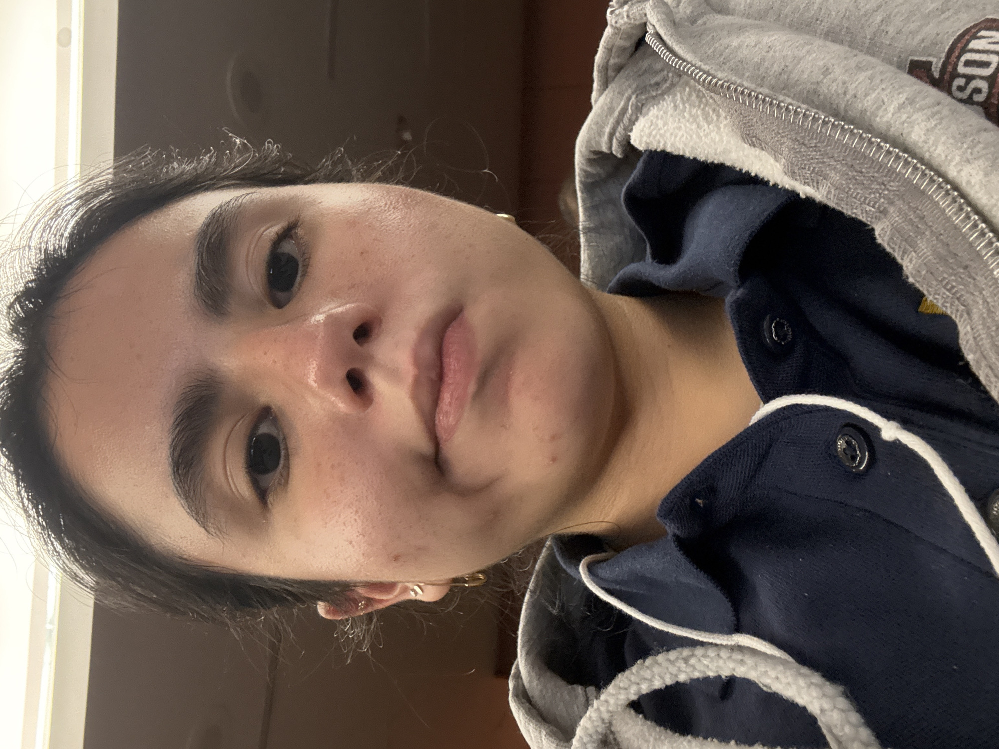 A close-up, low-angle portrait of a woman with dark hair. She has visible pores and some redness on her face. She is wearing a gray hooded sweatshirt over a navy blue collared shirt with white trim. A small gold earring is visible in her left ear. The background is blurred, suggesting an indoor setting with dark wood paneling. The image was taken from a close proximity, focusing on the woman's face and upper torso. No location in San Antonio, United States of America, can be determined from the provided image.