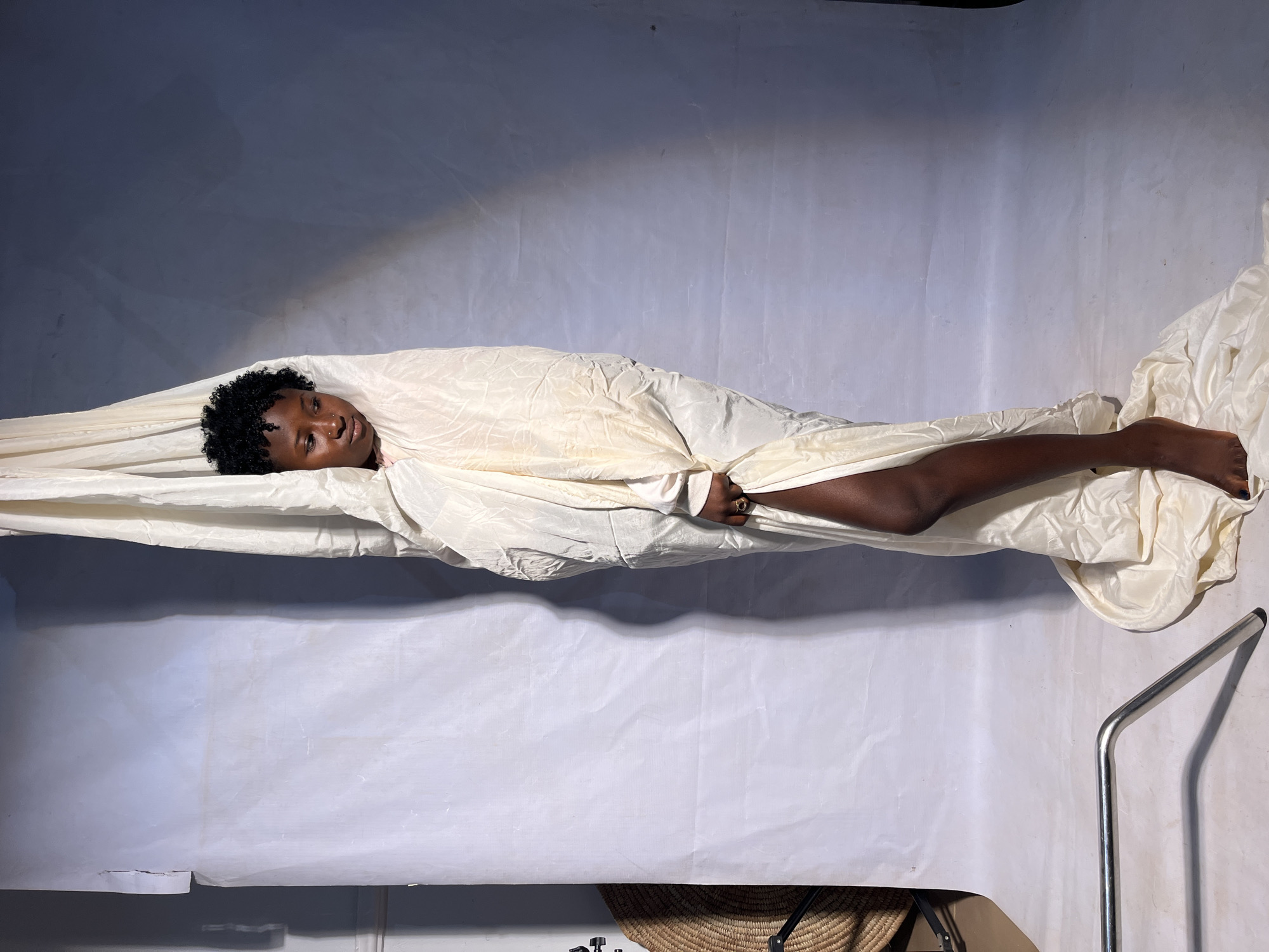 A young Black woman with short, curly hair is standing wrapped in a large, cream-colored piece of fabric. The fabric is draped from above, engulfing her body and pooling around her feet on the floor. One of her legs is exposed, with her foot bare. She is looking down and to her left. The background is a white backdrop with some shadowing. To the left of the woman, partially visible, is a wicker basket on a stand. In the foreground, a metallic silver stand is angled towards the viewer. The scene appears to be in a studio setting.
