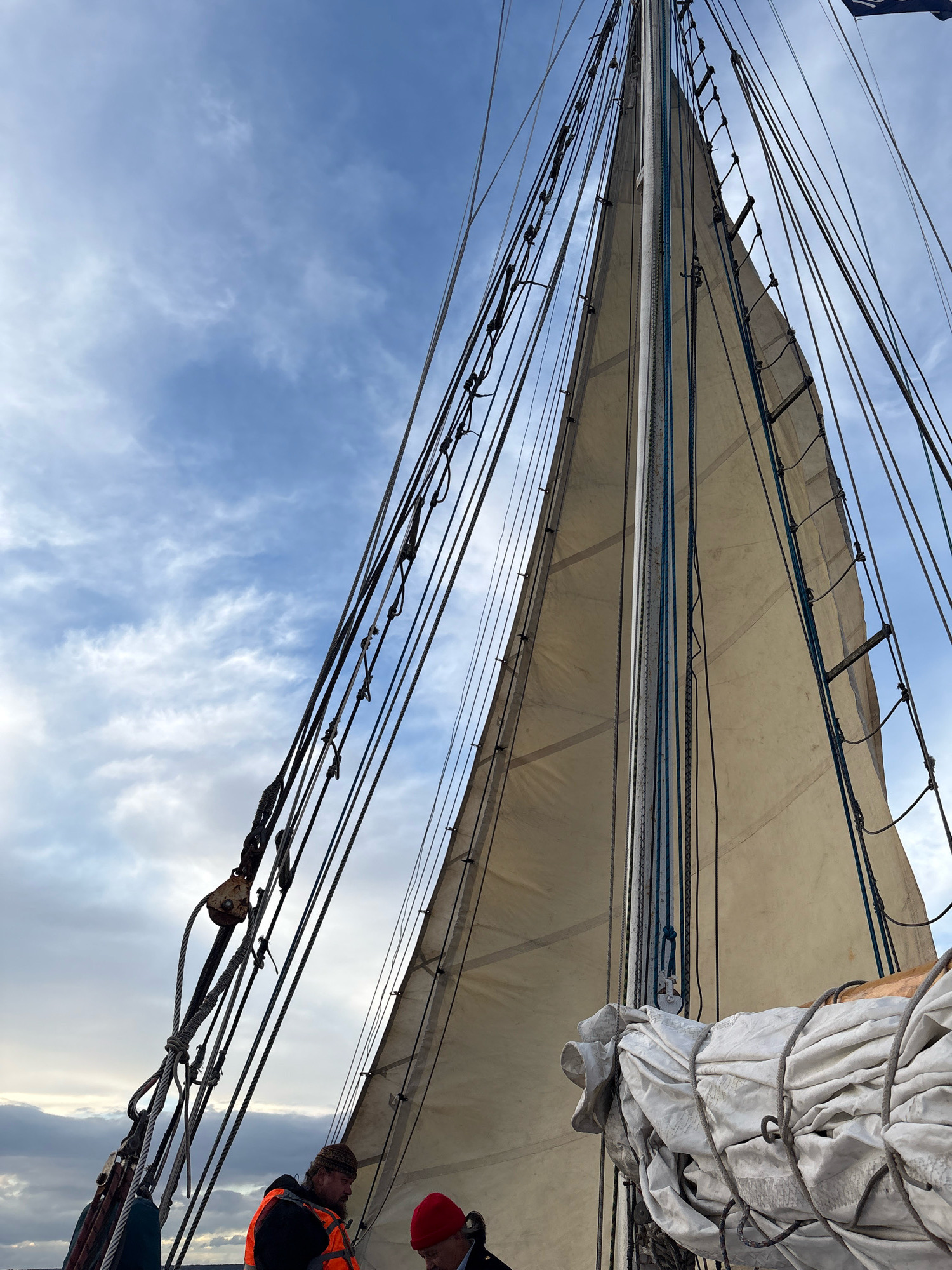 This image captures a close-up view of a sailboat's mast and sail against a cloudy sky, hinting at a maritime setting in Saint Petersburg, Russia. Two individuals are partially visible on deck, suggesting an active sailing experience. One person, wearing an orange safety vest and a patterned beanie, appears to be engaged in conversation or work, while the other, in a red beanie, looks down.
The sail, made of a light-colored fabric, is partially furled and secured with ropes. Intricate rigging, consisting of numerous ropes and pulleys, crisscrosses the frame, highlighting the complex mechanics of sailing. A large pulley system is prominent in the foreground on the left. The sky is a mix of blue and white clouds, indicating potentially variable weather conditions, possibly in the late afternoon or early morning given the soft light. The overall mood is one of preparation or ongoing activity on the water. There is no visible text in the image.