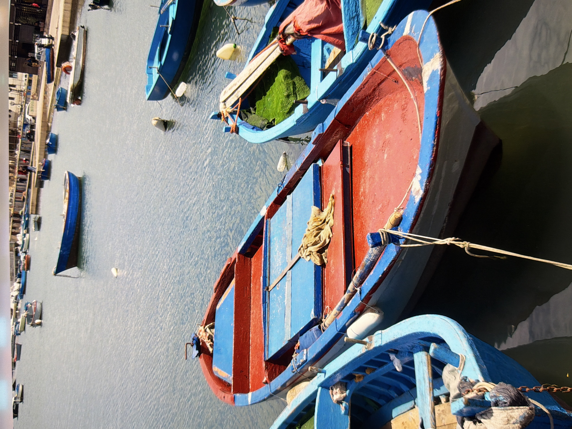 This media file depicts a cluster of colorful boats moored in a body of water, likely a harbor or canal. The boats are predominantly painted in vibrant red and blue hues, with some featuring white accents. Several of the boats are empty, while others appear to contain items like green material or folded fabric. In the background, a more distant collection of boats and a building or pier can be observed. The scene is bathed in natural light, suggesting it is daytime, and the water appears relatively calm. There are no explicit signs of human activity in the immediate foreground, but there are hints of people in the distant background. The overall atmosphere is one of a quiet, working harbor, possibly in a coastal town or city. The provided location context of Kyiv, Ukraine, adds a layer to the interpretation, though visual cues within the image itself are not specific enough to definitively confirm this location. The image is a still photograph with a slightly tilted perspective, giving a dynamic feel to the composition.