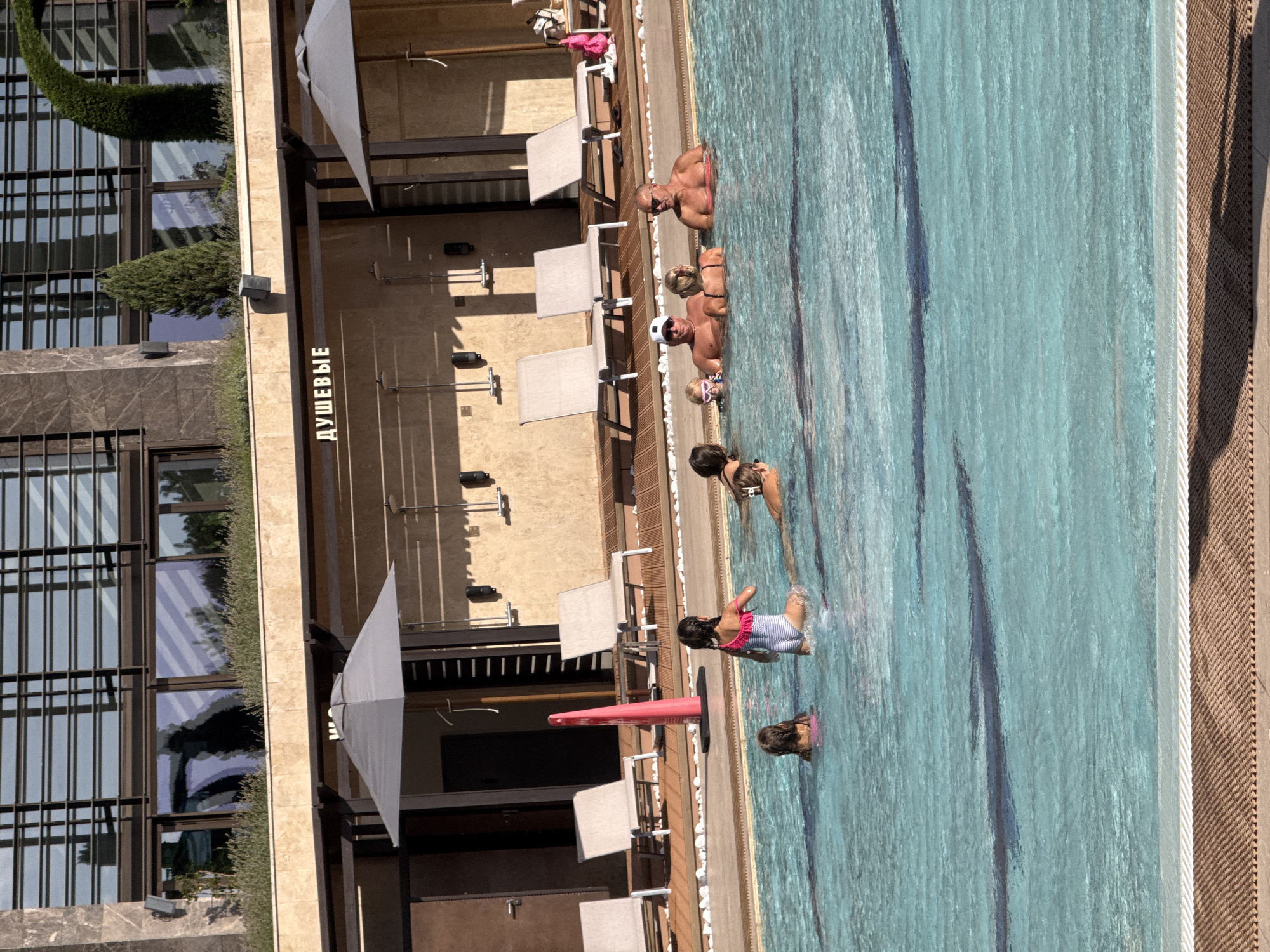 In an outdoor swimming pool area, likely in Simeiz, Ukraine, several people are enjoying the water on a sunny day. A group of adults and children are immersed in the turquoise water, with some adults standing and others interacting with the children. One young girl in a striped swimsuit is visible wading in the pool. Behind the pool, lounge chairs are arranged on a wooden deck, some shaded by large umbrellas. A building with modern architecture and large windows forms the backdrop, with the word "ДУШЕВЫЕ" (showers) visible above the pool area, indicating the presence of shower facilities. The environment appears to be a resort or a similar recreational facility.