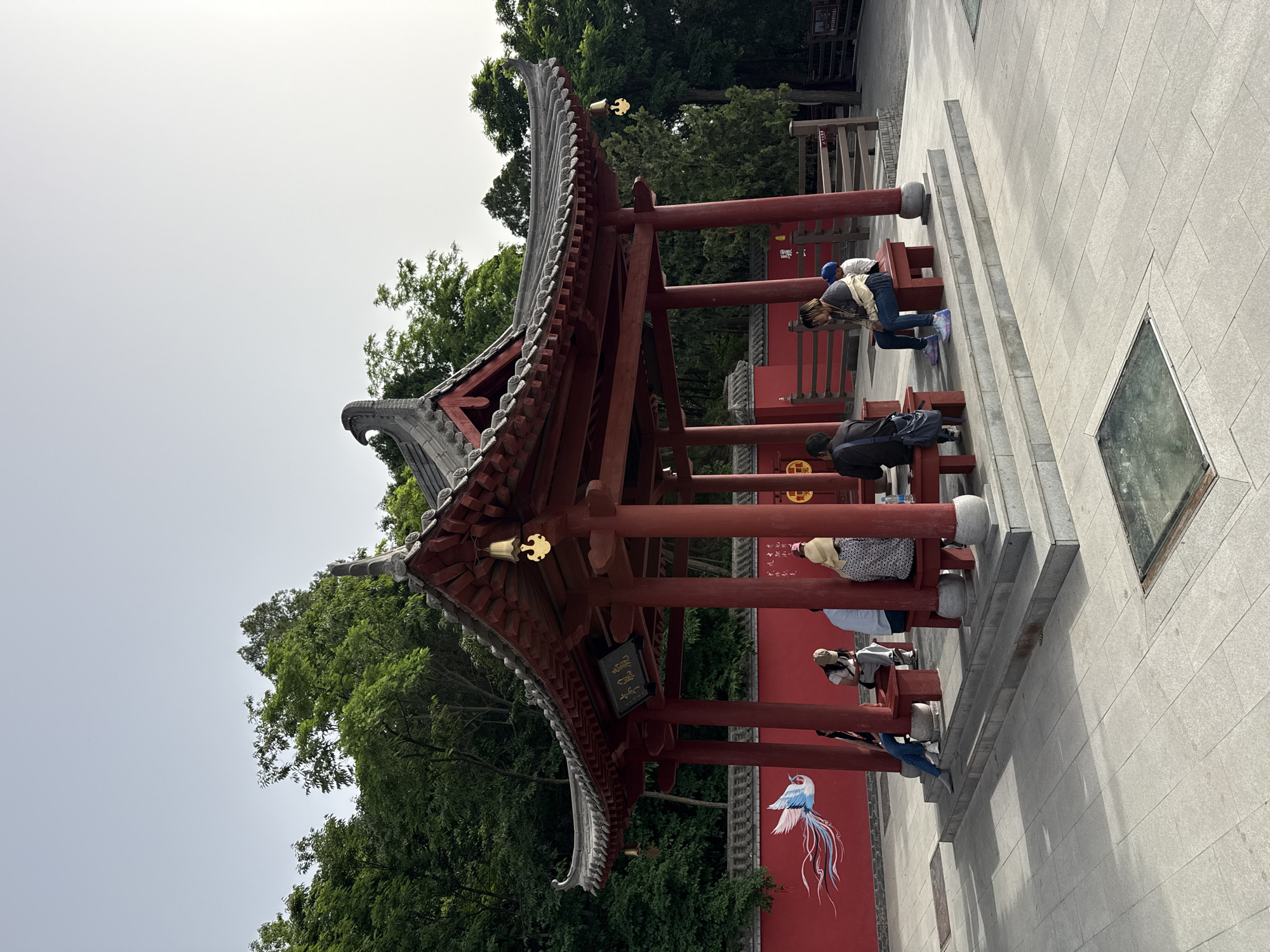 The image depicts a red pavilion structure in Lintong District, China, with people sitting on benches inside. The pavilion features a traditional Chinese roof design with gray tiles and ornate eaves. The surrounding environment includes trees and a red wall with a mural of a bird. The ground is paved with stone tiles. The sky is overcast.