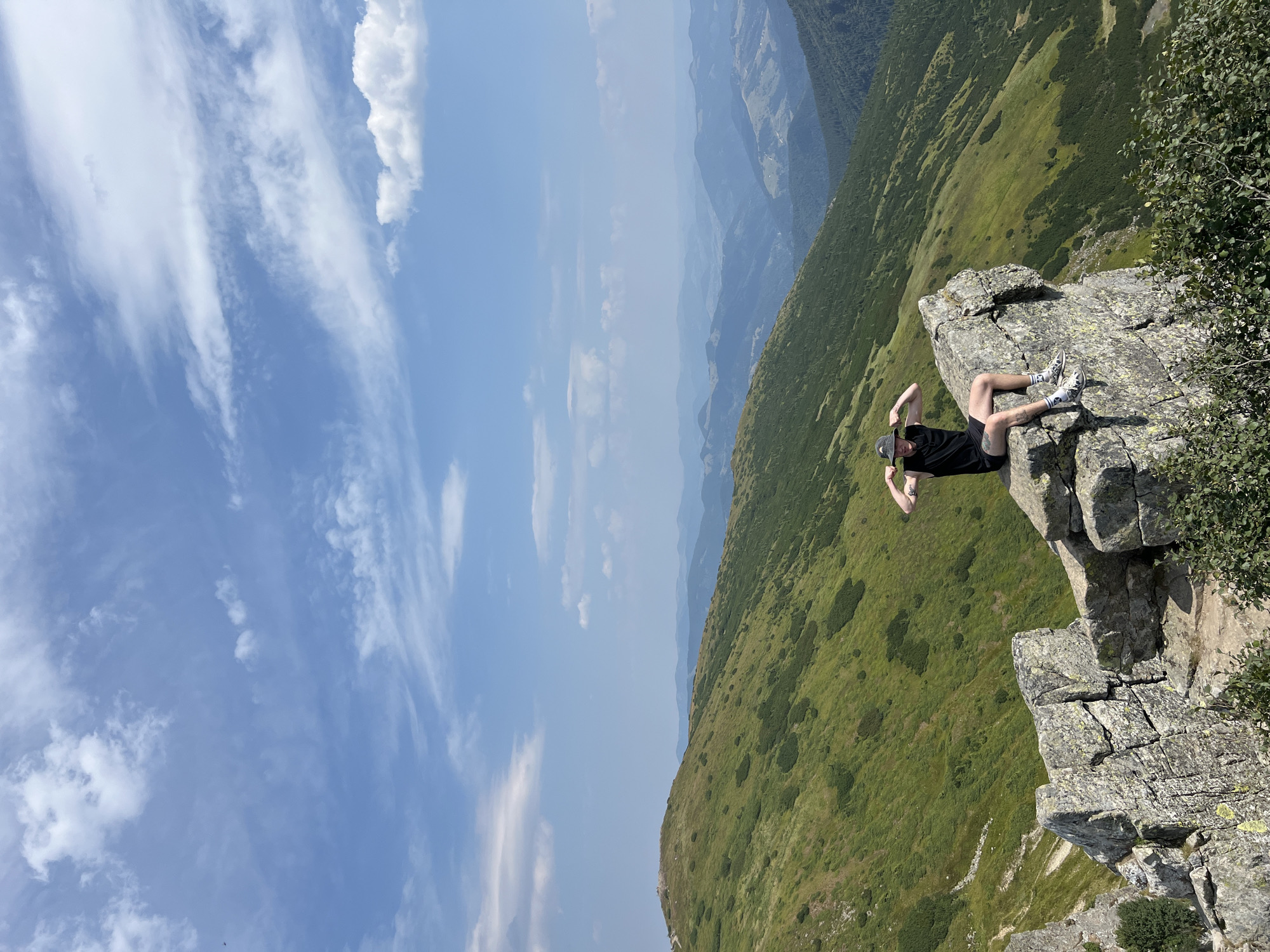 A young person wearing a black tank top, black shorts, a bucket hat, and patterned socks sits on a large rock formation. The person is flexing their biceps and looking towards the camera. They are positioned on the edge of a cliff with a steep, green, mountainous landscape stretching out behind them, leading to layers of hazy blue mountains in the distance under a bright, partly cloudy sky. The location is a mountain peak in the Carpathian Mountains, near the town of Vorokhta, Ukraine, a popular destination for hiking and outdoor recreation.