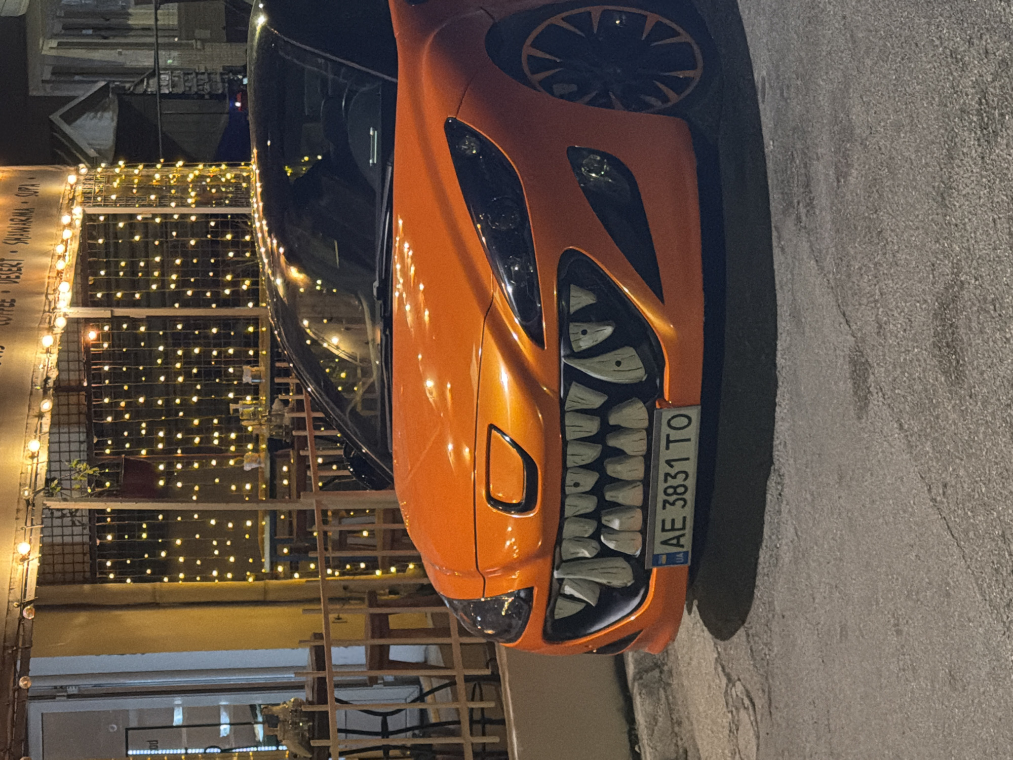 An orange car with a custom grille designed to resemble large teeth is parked on a street at night. The car has its headlights on, illuminating the road and the sidewalk. In the background, a building with string lights and an outdoor seating area is visible. A sign above the outdoor seating area displays text including "COFFEE," "DESSERT," "SHAWARMA," and "SOUP." The license plate of the car reads "AE 3831 TO." The scene appears to be in a city, likely in a commercial area with restaurants or cafes. The overall impression is of a car modified for a festive or novelty purpose, parked in an urban environment at night.