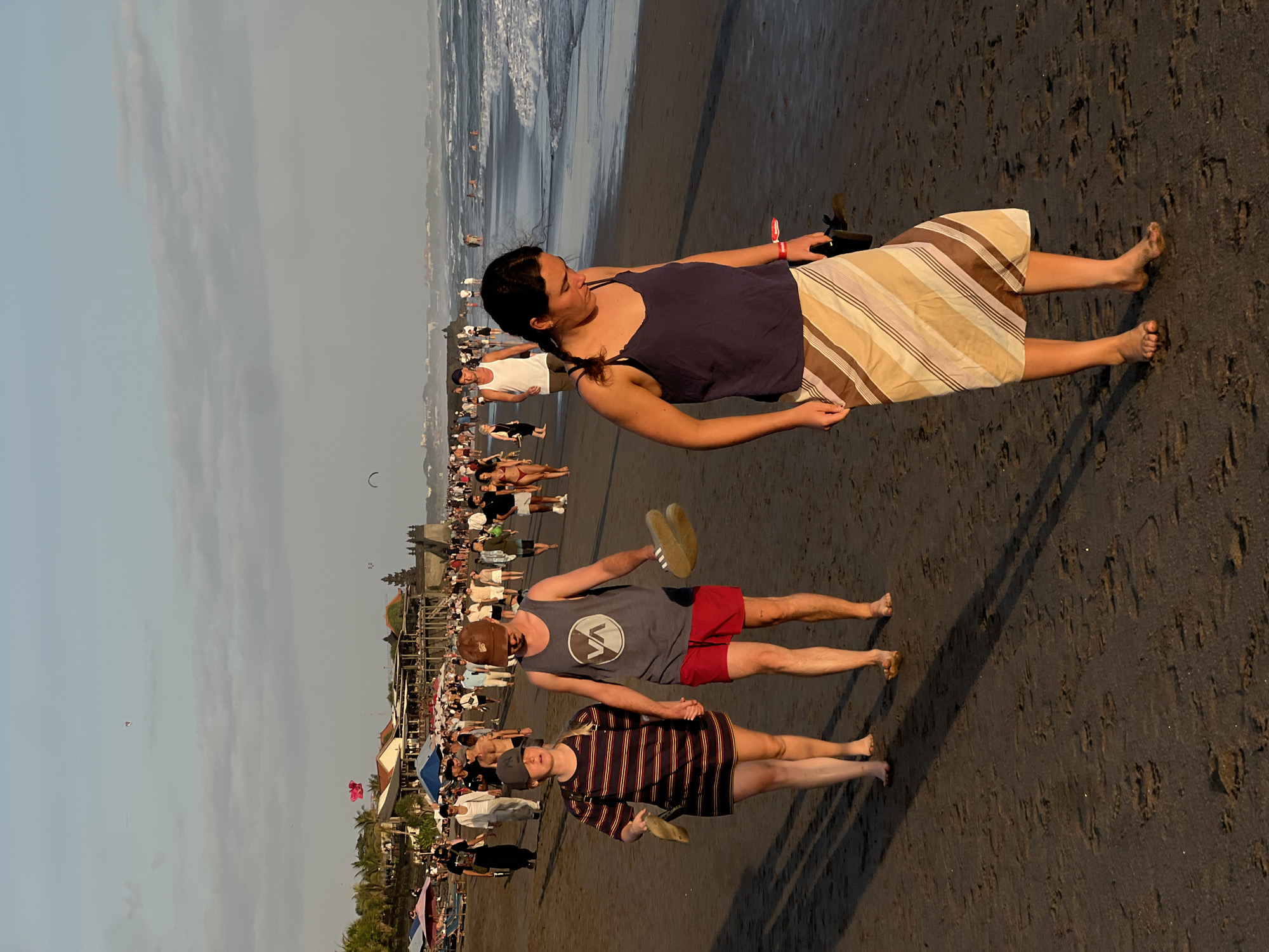 In North Kuta, Indonesia, a beach scene unfolds at sunset. The tide is low, with gentle waves lapping the shore. Numerous people are present, some walking along the beach, others in the water, and some gathered around beach umbrellas. The sand is dark and wet, reflecting the warm hues of the setting sun.
In the foreground, three individuals are walking towards the viewer. A woman in a blue tank top and a striped skirt is on the right, holding a pair of flip-flops. To her left, a couple walks hand-in-hand. The woman wears a striped dress and carries flip-flops. The man wears a grey tank top with a logo and red shorts.
In the background, a line of buildings with thatched roofs runs along the coastline, dotted with umbrellas and beach chairs. Further out, the ocean stretches to the horizon, where the sky transitions from a soft blue to warmer tones. Several kites are visible in the air, adding to the festive atmosphere.