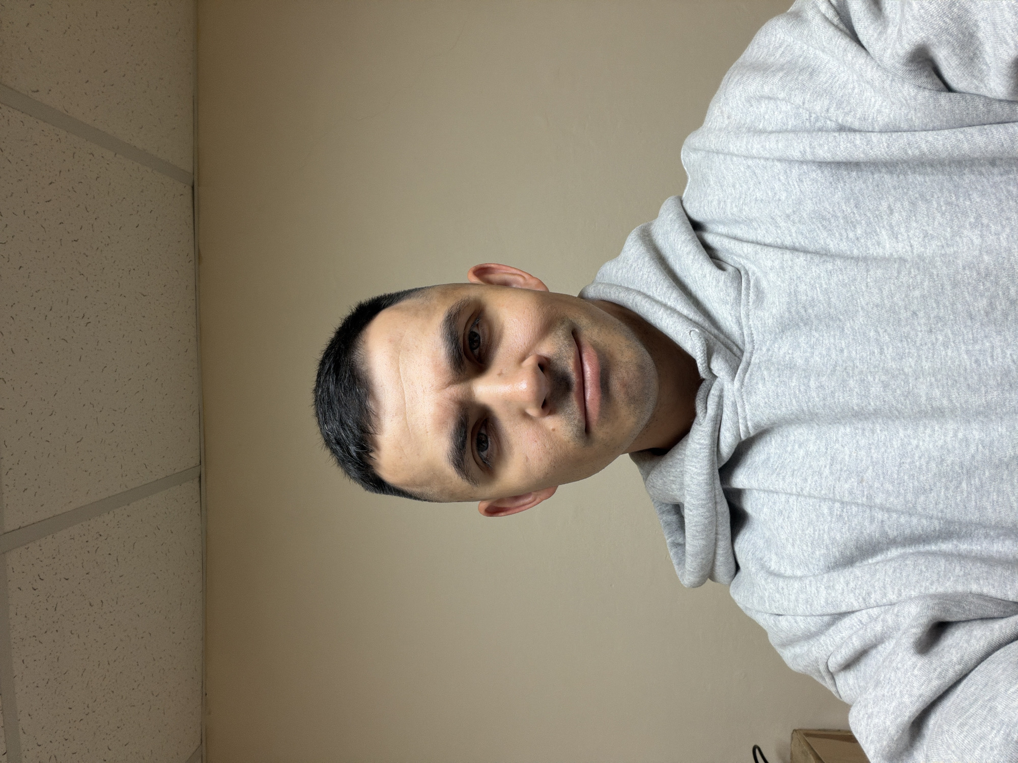 A man with short black hair and fair skin is wearing a grey hooded sweatshirt. He appears to be indoors, with a beige wall and a ceiling with acoustic tiles visible in the background. The location is not discernable as Izhevsk, Russia, or anywhere else based on the provided image.