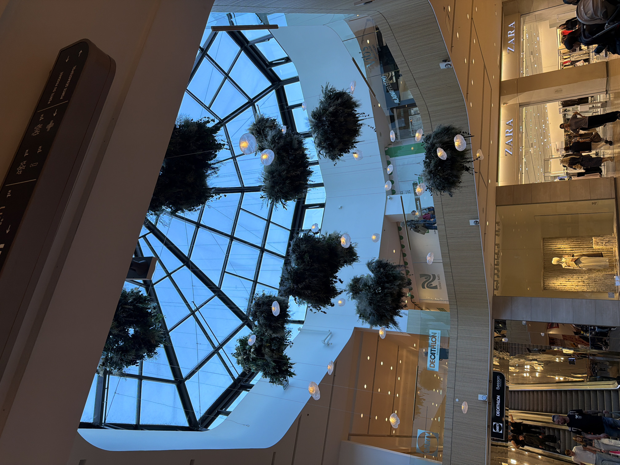 The image depicts the interior of a modern, multi-level shopping mall, likely located in Strasbourg, France, as indicated by the "DECATHLON STRASBOURG - LES HALLES" signage.
The main subject is the mall's architecture and decorations. A large, geometric glass skylight forms the ceiling, allowing natural light to filter in, revealing a light blue sky, suggesting it is daytime. Suspended throughout the central atrium are numerous large, spherical clusters of green foliage, possibly artificial plants or dried elements, intertwined with small, glowing white orb-shaped lights, contributing to an elegant and natural aesthetic.
Several people, appearing to be shoppers, are visible on different levels of the mall, walking or browsing. Stores are visible on multiple floors, with prominent signage for "ZARA" on the upper right side. Other visible retailers include "DECATHLON" (along with "Besson" and "DARTY" nearby), "Golden Nails," "ALAINAFFLELOU," "CACHE CACHE," and "BONOBO."
A dark grey directional sign on the left wall provides information in French, reading "PORTE DU PASSAGE" with an upward arrow and "PORTE SEBASTOPOL" with a left arrow. It also features icons for food/drink services and level indicators like "7ea" and "7eb." The overall scene portrays a bright, spacious, and active commercial environment.