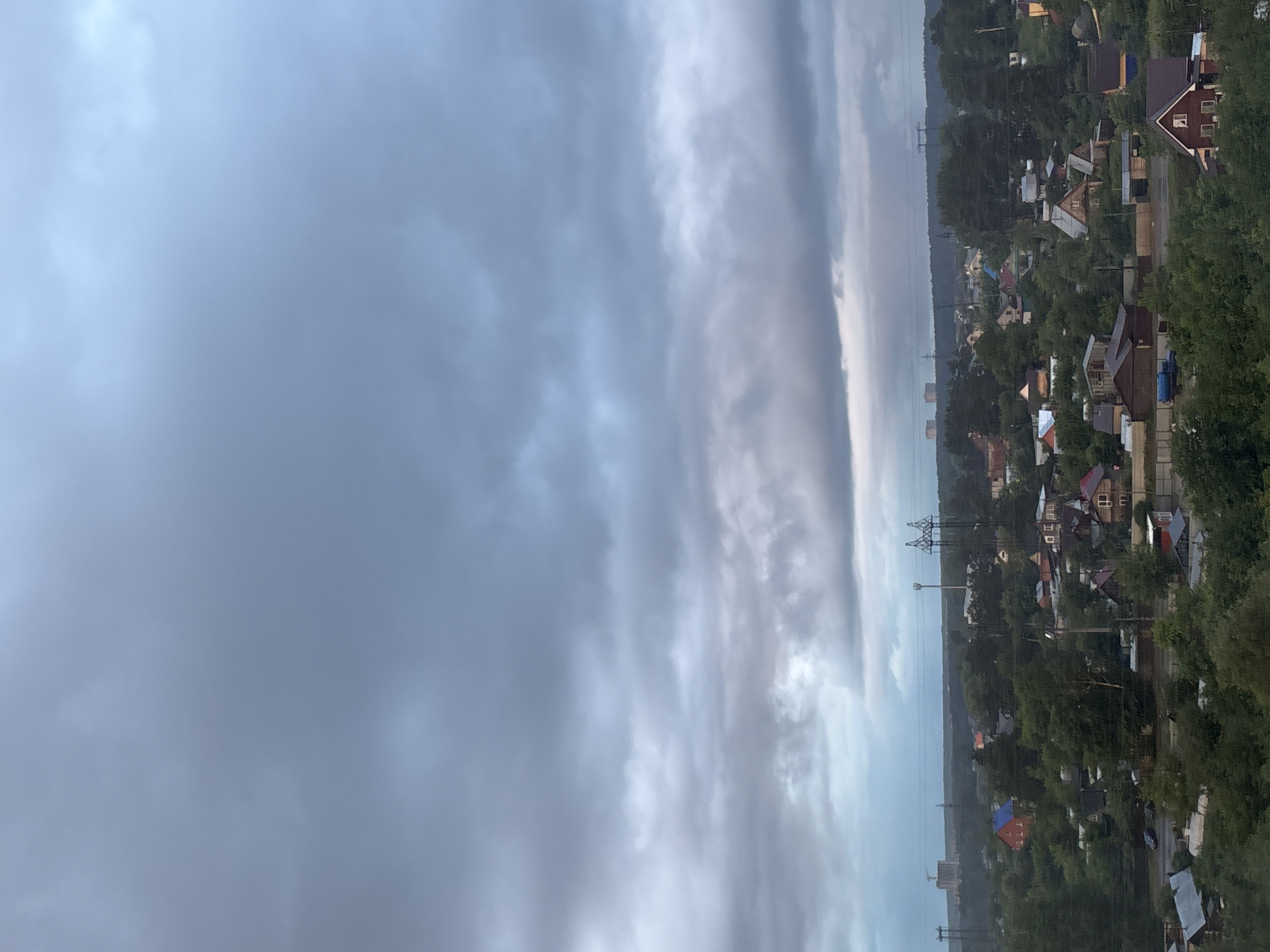 The image captures a broad, somewhat elevated view of a settlement, likely a village or suburban area, under a heavily overcast sky. The scene depicts numerous houses of varying sizes and colors, predominantly with sloped roofs, nestled closely among a dense canopy of green trees. Some houses are painted in light colors, while others feature darker wood or brick facades. A road or path appears to weave through the settlement.
Prominent features in the landscape are several large power line towers and associated electrical lines that traverse the area, indicating utility infrastructure. The sky occupies a significant portion of the image, filled with elongated, dense clouds that range from light grey to darker, ominous shades. The overall lighting is diffused, suggesting a cloudy day, possibly late afternoon or early evening, or simply a fully overcast sky. There are no people visible, and no specific activities are taking place. The setting is consistent with a residential area in a natural, tree-filled environment. Based on the provided context, the location is Chaikovsky, Russia. There is no visible text in the image.