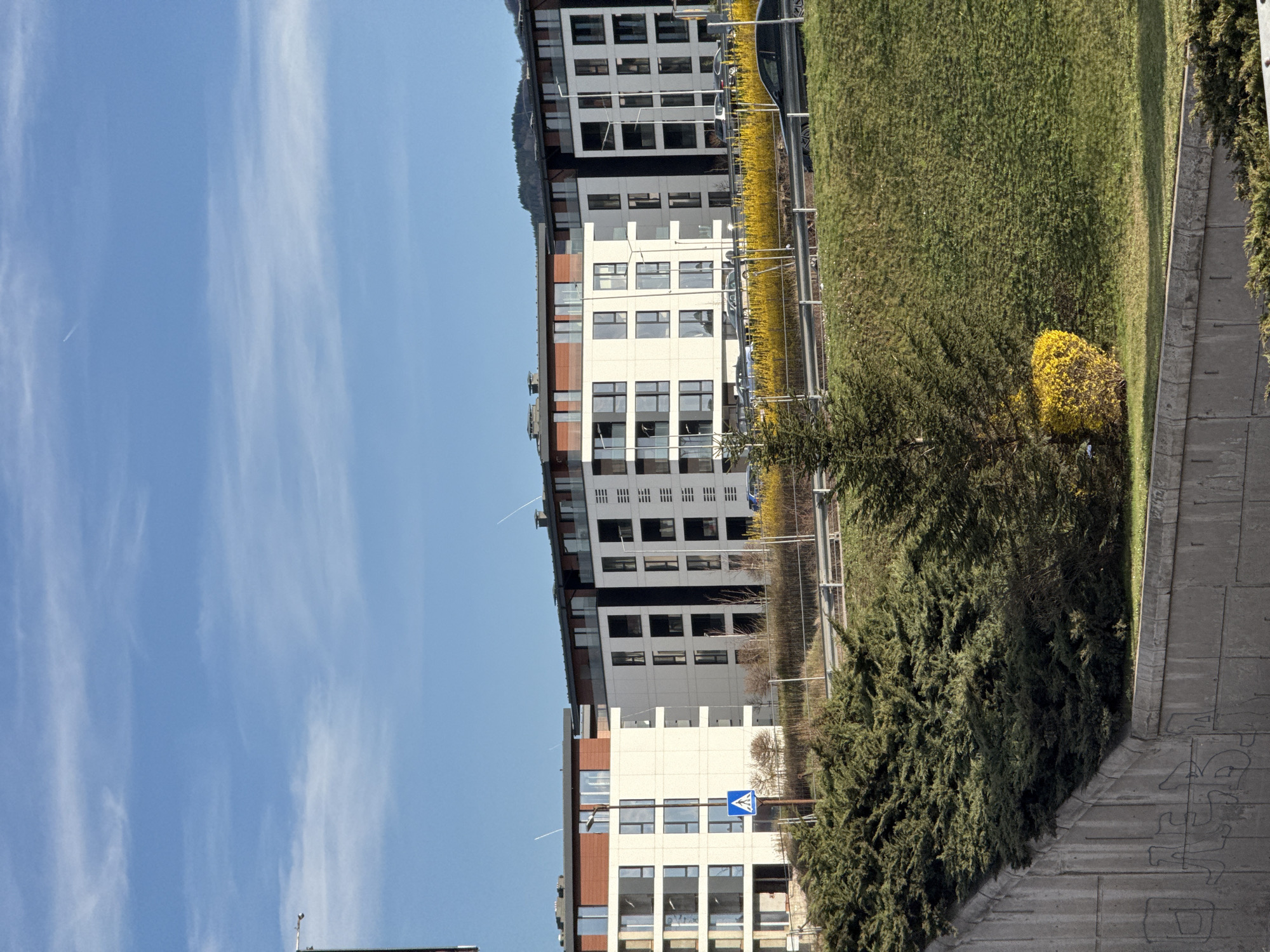 The image shows a view in Sofia, Bulgaria, with a multi-story building dominating the scene. The building has a modern architectural design featuring a white facade with dark window frames and brown accents. A pedestrian crossing sign is visible in front of the building. The building is surrounded by greenery, including grass, bushes, and a yellow flowering bush. A fence runs along the edge of the grass. A blue sky with light, wispy clouds is visible above the building. A concrete walkway is visible in the foreground. The overall scene appears to be a sunny, urban environment in Sofia, Bulgaria.