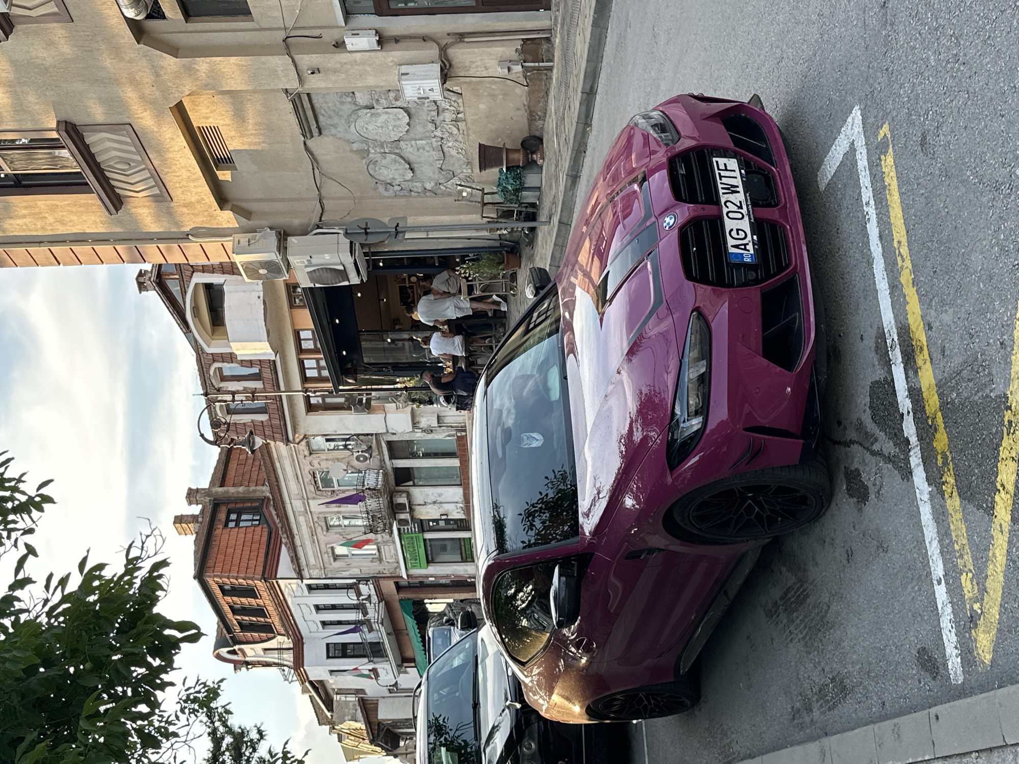 In Veliko Tarnovo, Bulgaria, a striking purple BMW M4 coupe is parked on a cobblestone street, angled towards the viewer. The car features black racing stripes on the hood and has a Bulgarian license plate reading "AG 02 WTF." Behind the car, a black SUV is partially visible. The street is lined with multi-story buildings exhibiting eclectic architecture, with some facades displaying ornate details and balconies adorned with Bulgarian flags. One building features an establishment with outdoor seating, where several individuals are visible. The scene is illuminated by natural light, casting shadows and highlighting the textures of the buildings and street. The overall atmosphere suggests a vibrant urban setting.
